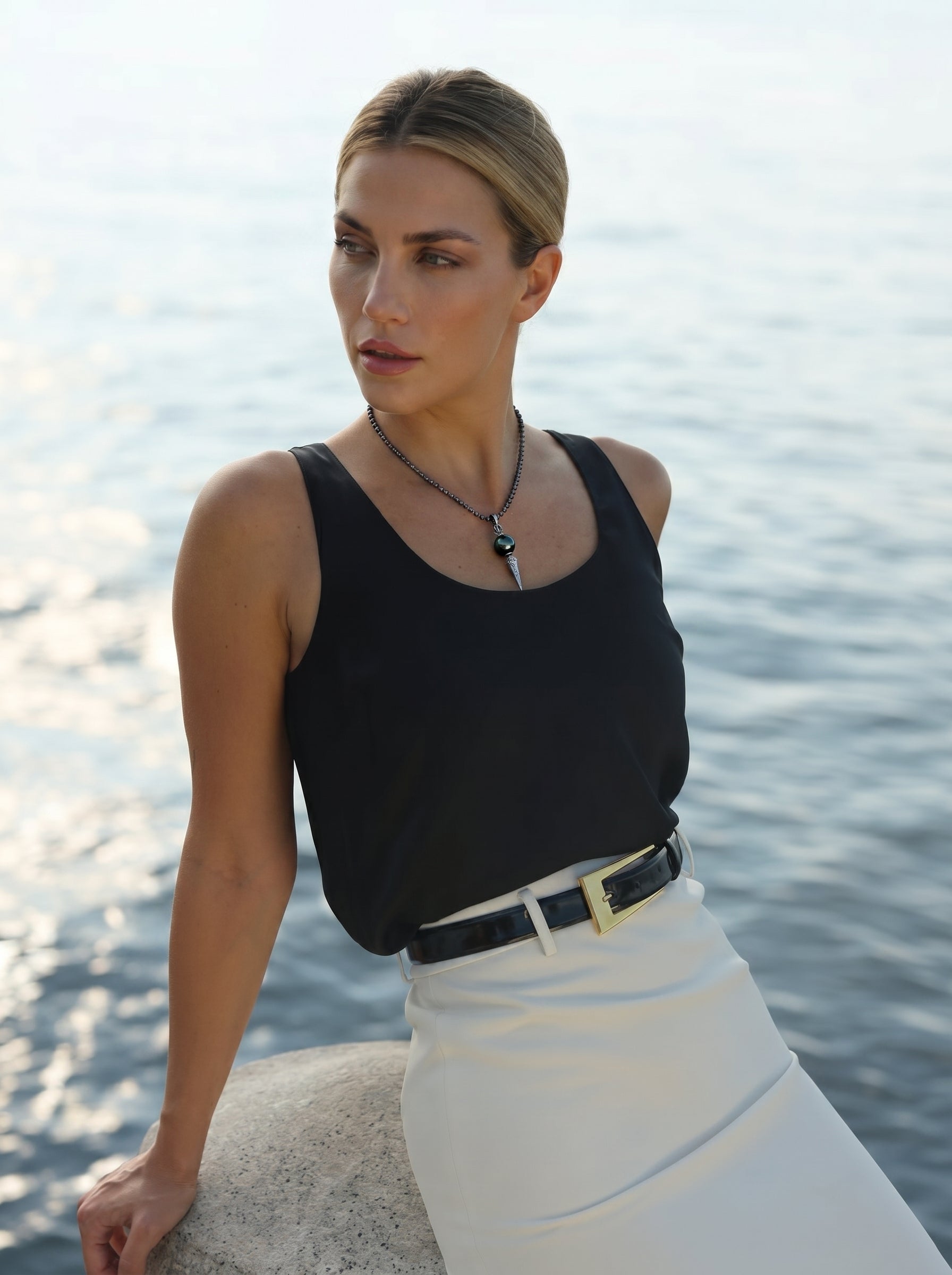 A woman wearing the Abyss Spire Tahitian Pearl Phurba necklace with a black tank top and white skirt, posing outdoors by the water. @lifestyle #lifestyle