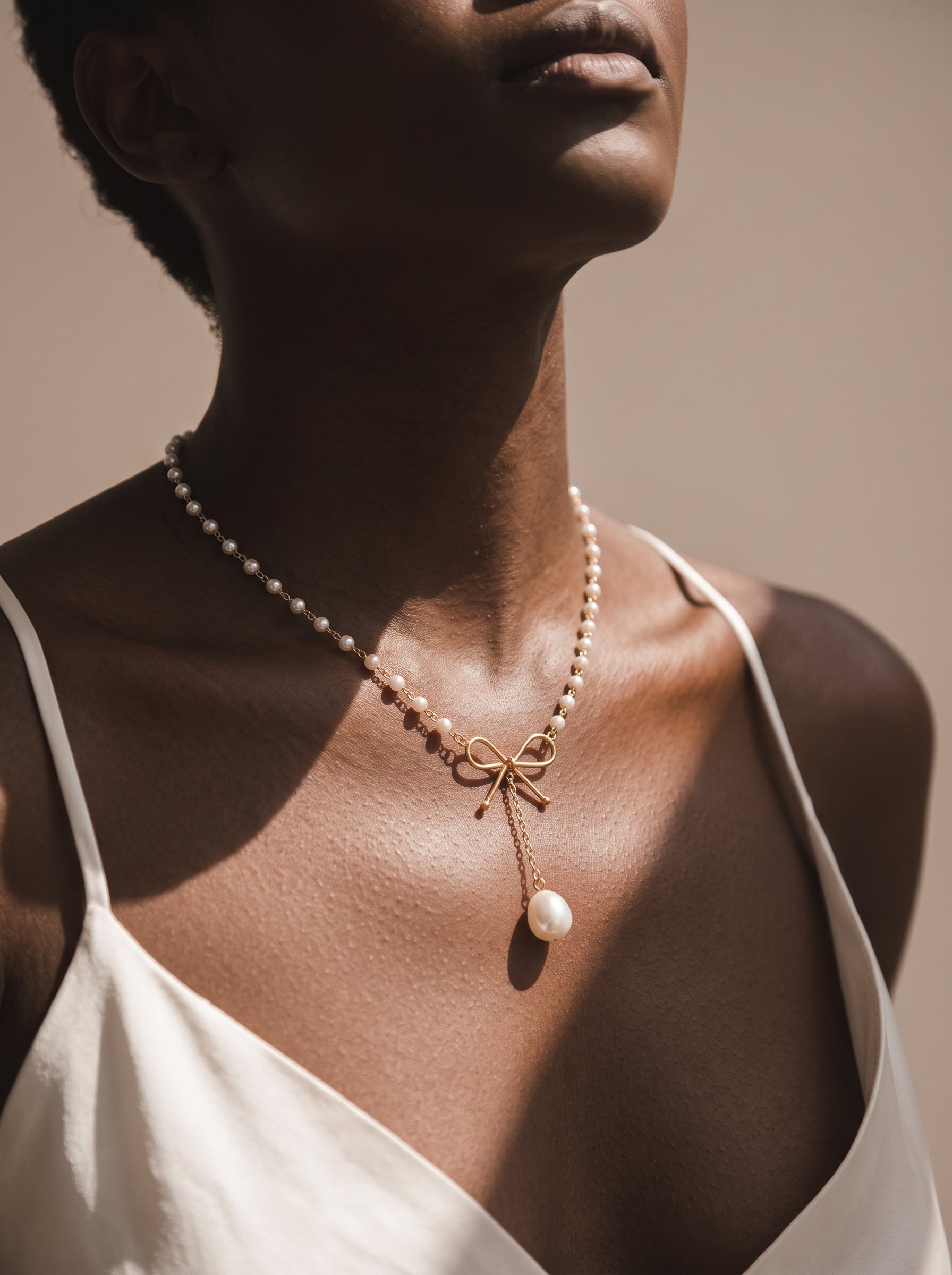 A model wears a delicate gold-tone necklace featuring a beaded chain of small imitation pearls, a central bow accent, and a dangling larger imitation pearl pendant. @model