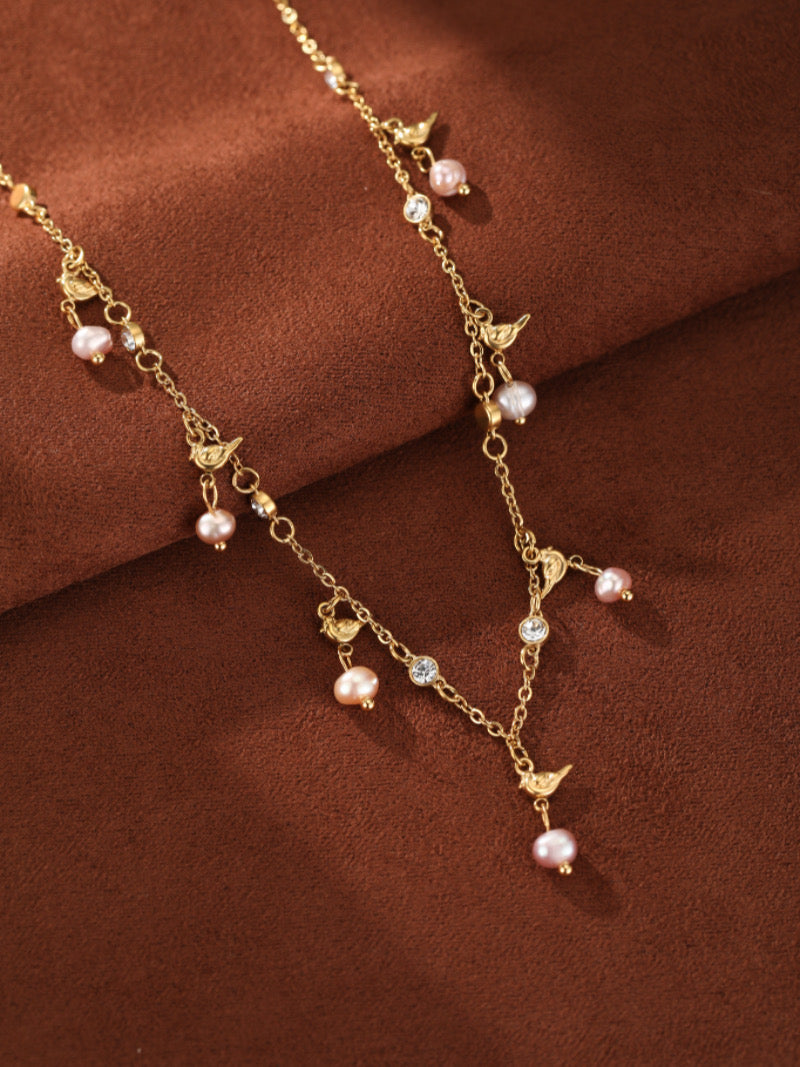 Un délicat collier flottant ton or présentant de multiples petites perles roses d'eau douce, de charmantes breloques oiseau, et des accents en cristal transparent sur fond texturé marron.
