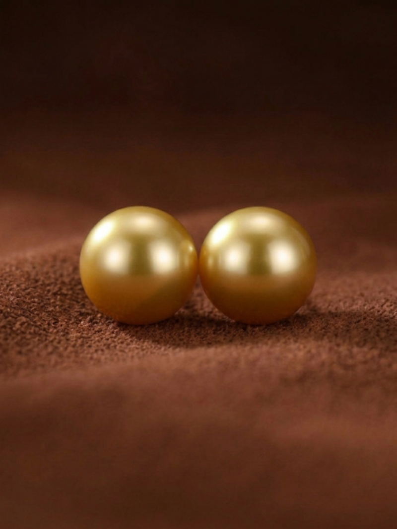 Two lustrous, perfectly round golden freshwater pearl stud earrings on a textured brown background.