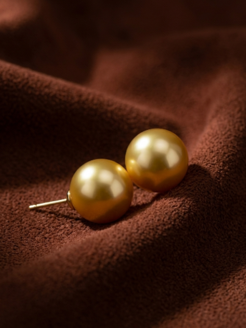 Golden freshwater pearl stud earrings with a lustrous sheen, presented on a textured brown fabric surface.