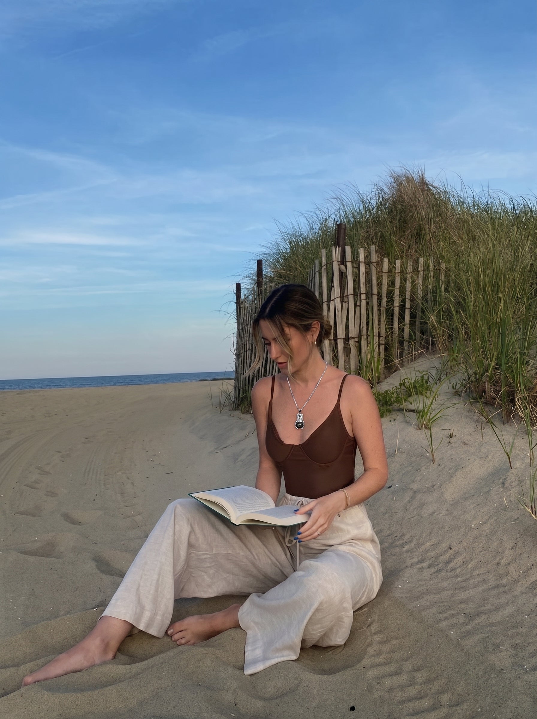 Une femme est assise sur une plage de sable en train de lire un livre, portant le collier pendentif tiki Ancestral en perle de Tahiti présentant un pendentif tiki argenté avec une perle noire. @lifestyle