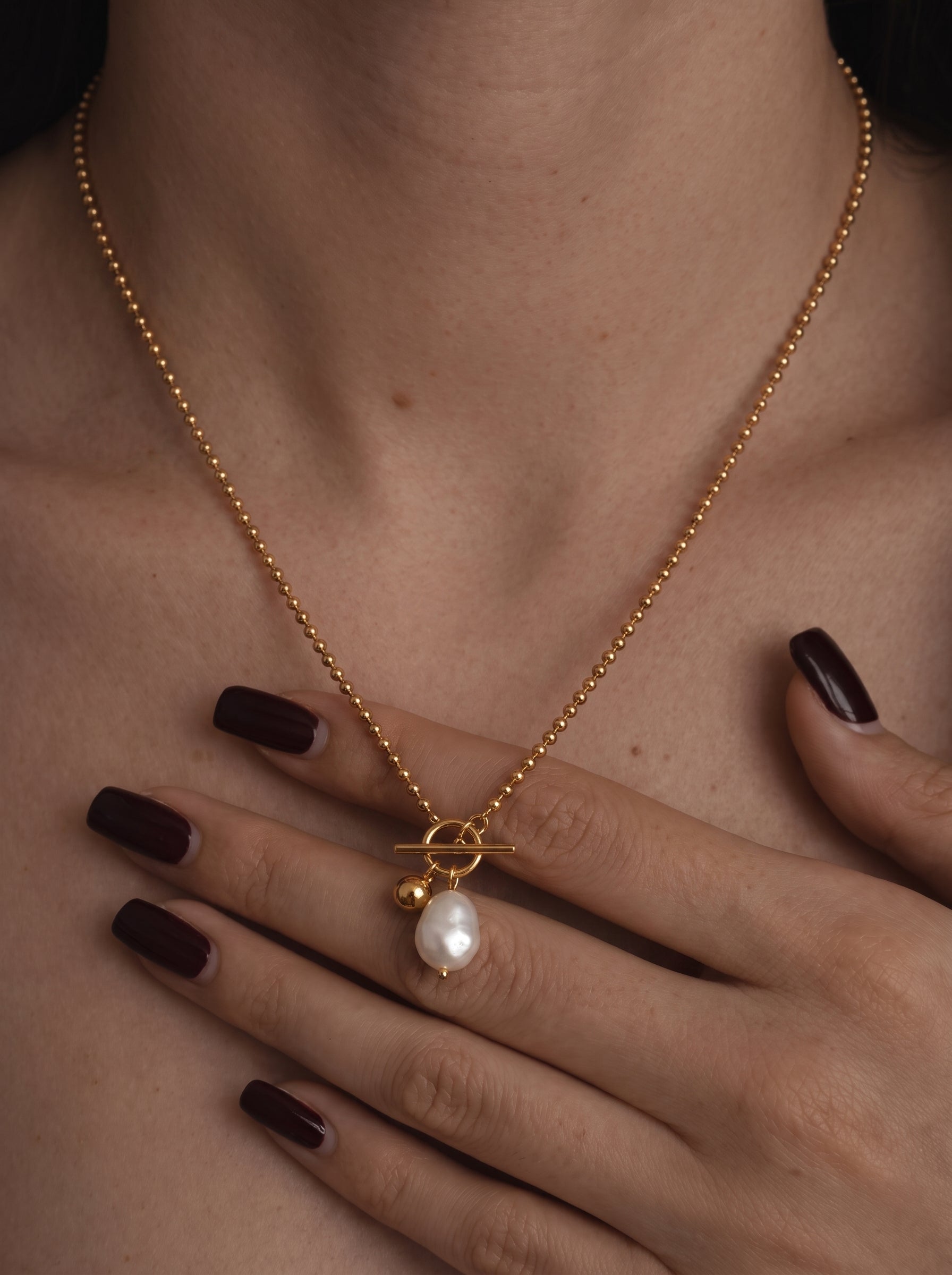 Close-up of a gold-toned ball chain necklace with a chunky pendant featuring a white drop freshwater pearl and a small gold bead, worn by a model. @model