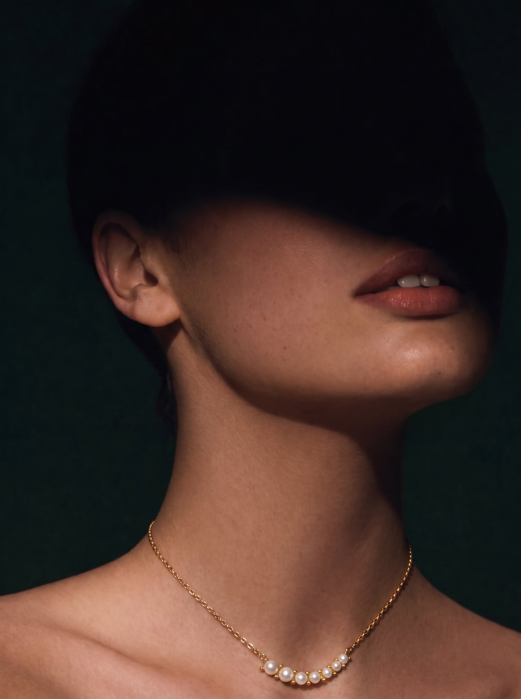 A close-up shot of a model wearing the Arcadia Freshwater Pearl Curved Bar Necklace, featuring a delicate gold chain with a curved bar adorned with seven small, luminous white pearls. The model's face is partially shadowed, with emphasis on her lips and neck where the necklace rests. @model
