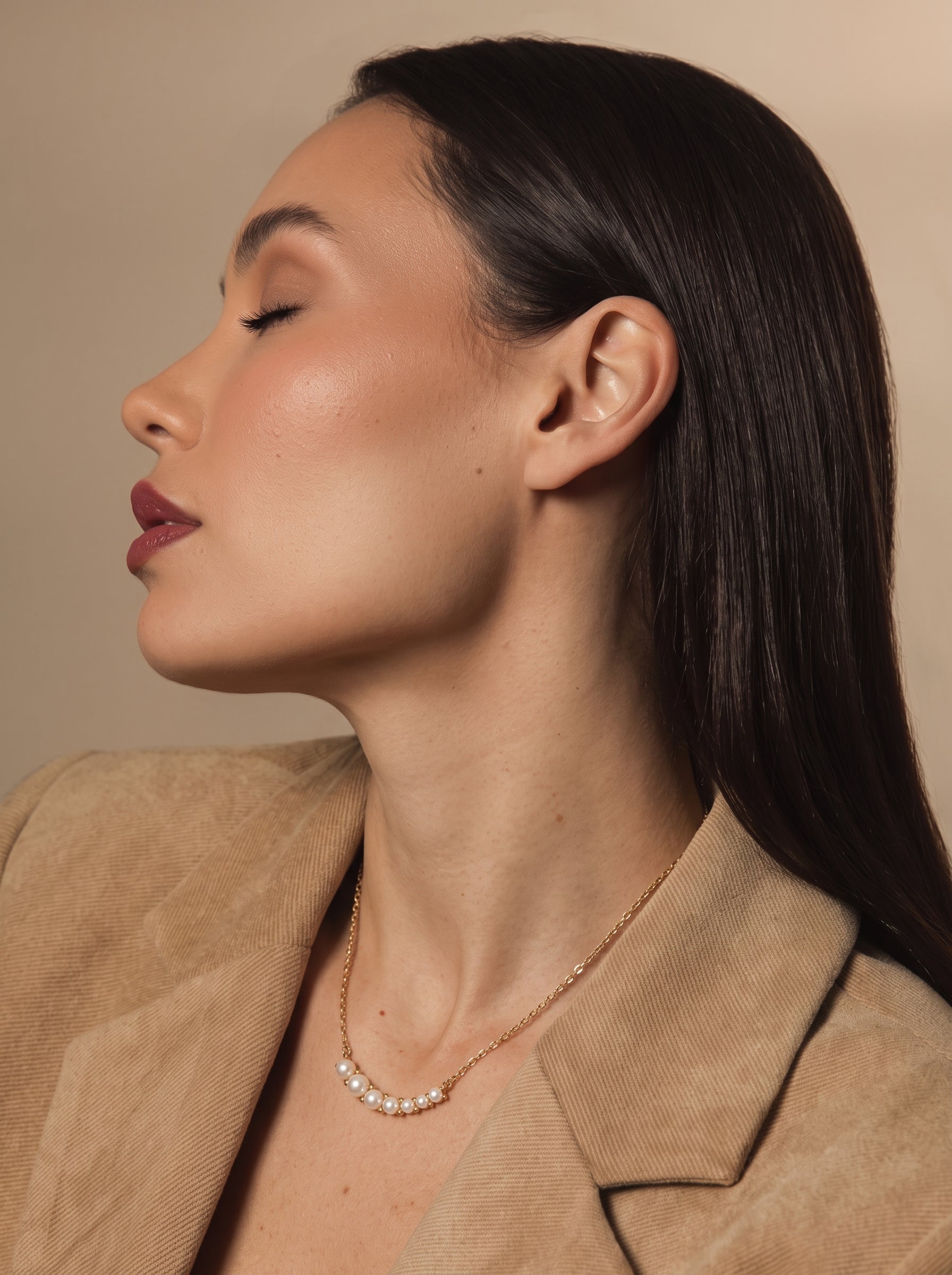 Model wearing the Arcadia Freshwater Pearl Curved Bar Necklace, featuring a delicate gold chain and a row of seven small, luminous white pearls arranged in a gentle curve across her décolletage. She is wearing a beige blazer and her eyes are closed. @lifestyle