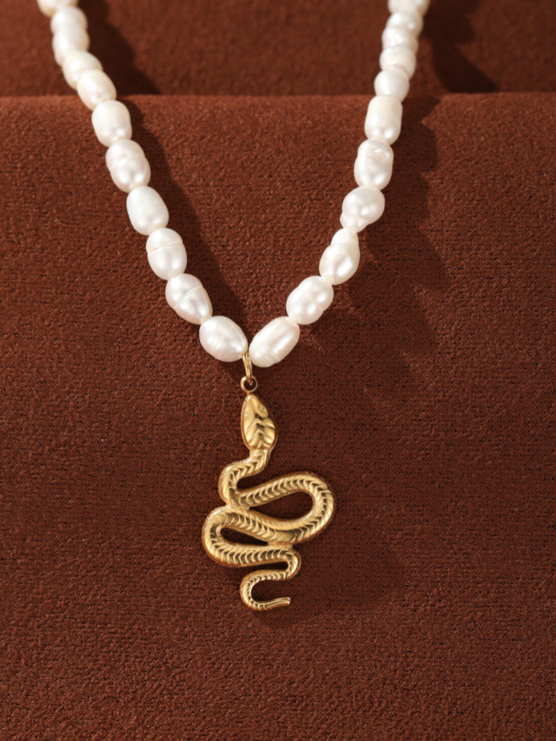 Gros plan du collier pendentif perlé serpent Ascendant en perle d'eau douce, présentant un rang de lustrées perles graines blanches et un pendentif serpent détaillé ton or sur fond texturé marron.