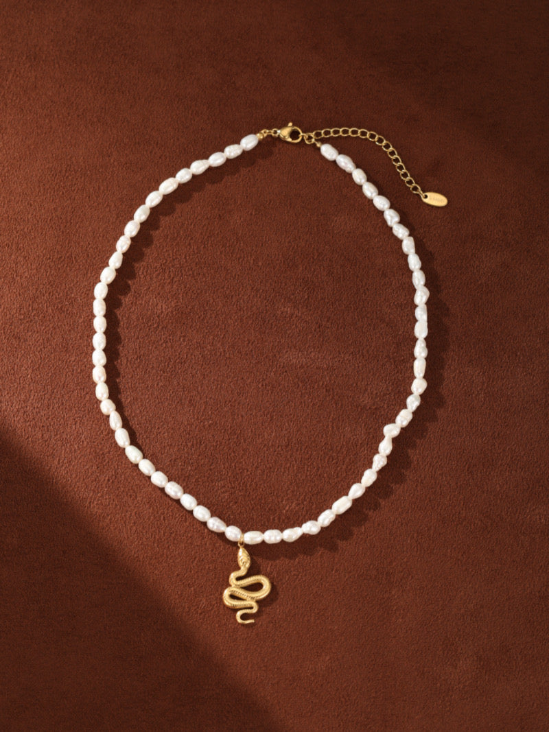 Collier pendentif perlé serpent Ascendant en perle d'eau douce présentant un pendentif serpent ton or sur un fil de perles blanches irrégulières d'eau douce contre un fond marron.