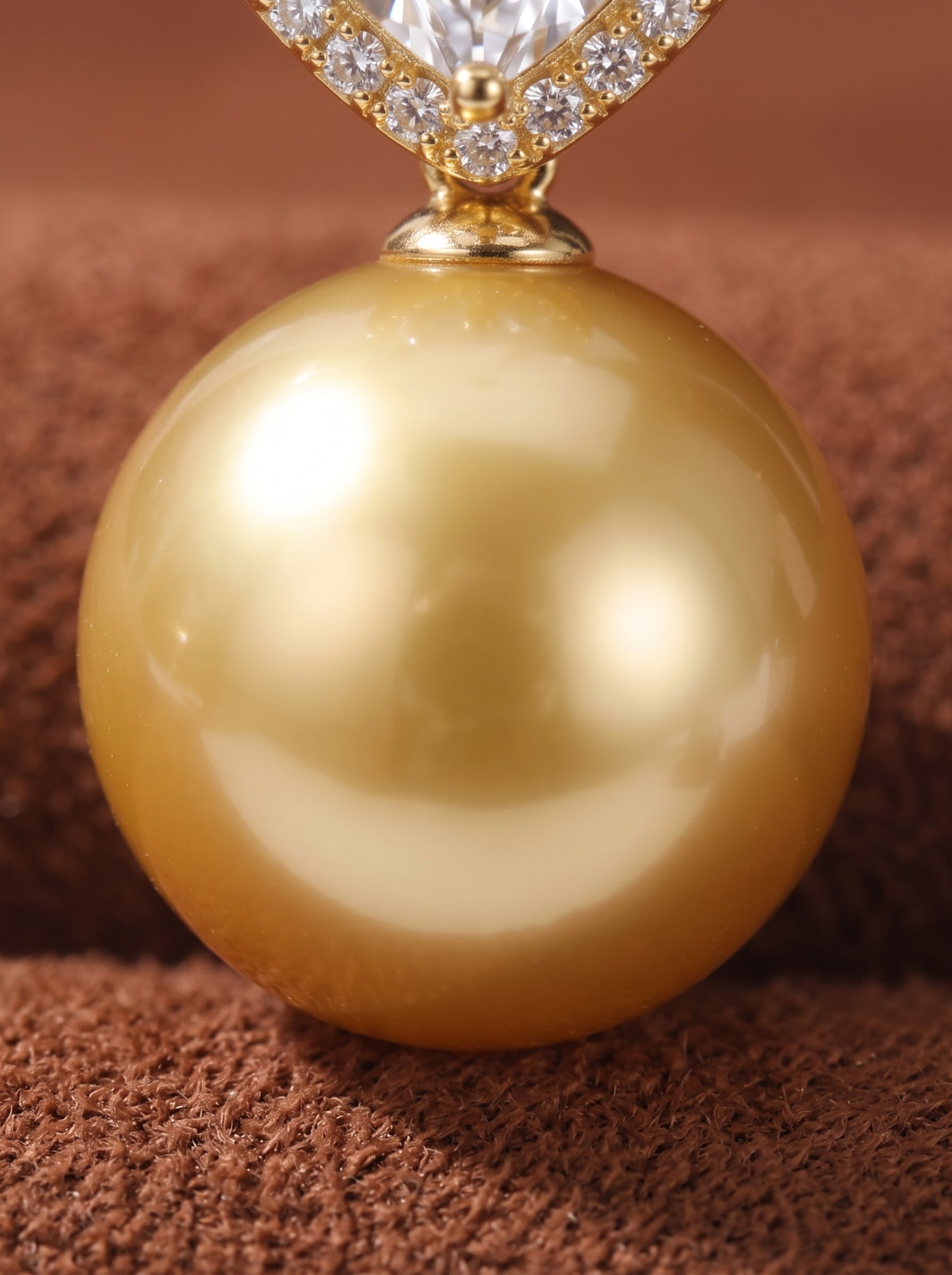 Close-up of a large, lustrous 15mm golden South Sea pearl pendant with a square bail adorned with small clear stones, resting on a textured brown surface. @pearl
