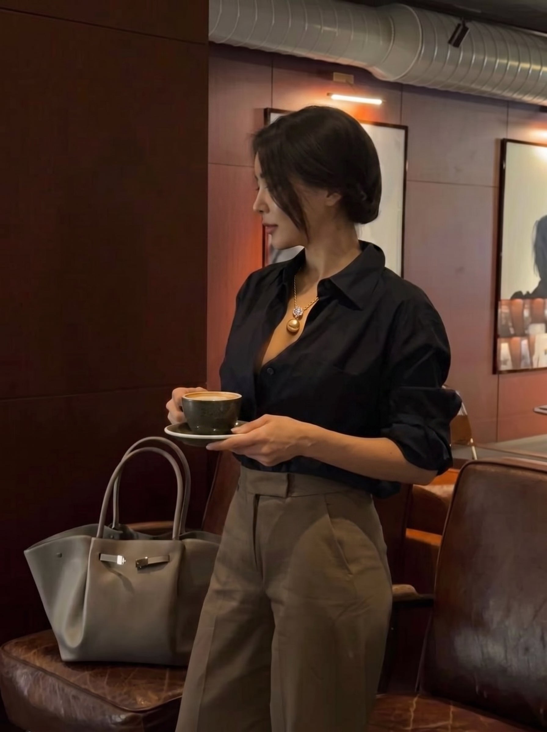 A woman in a black shirt and brown pants holds a cup of coffee while wearing the Aurelia South Sea Pearl Square Single Pendant Necklace, featuring a large golden pearl suspended from a square bail with accent stones. A stylish grey tote bag sits beside her. @lifestyle