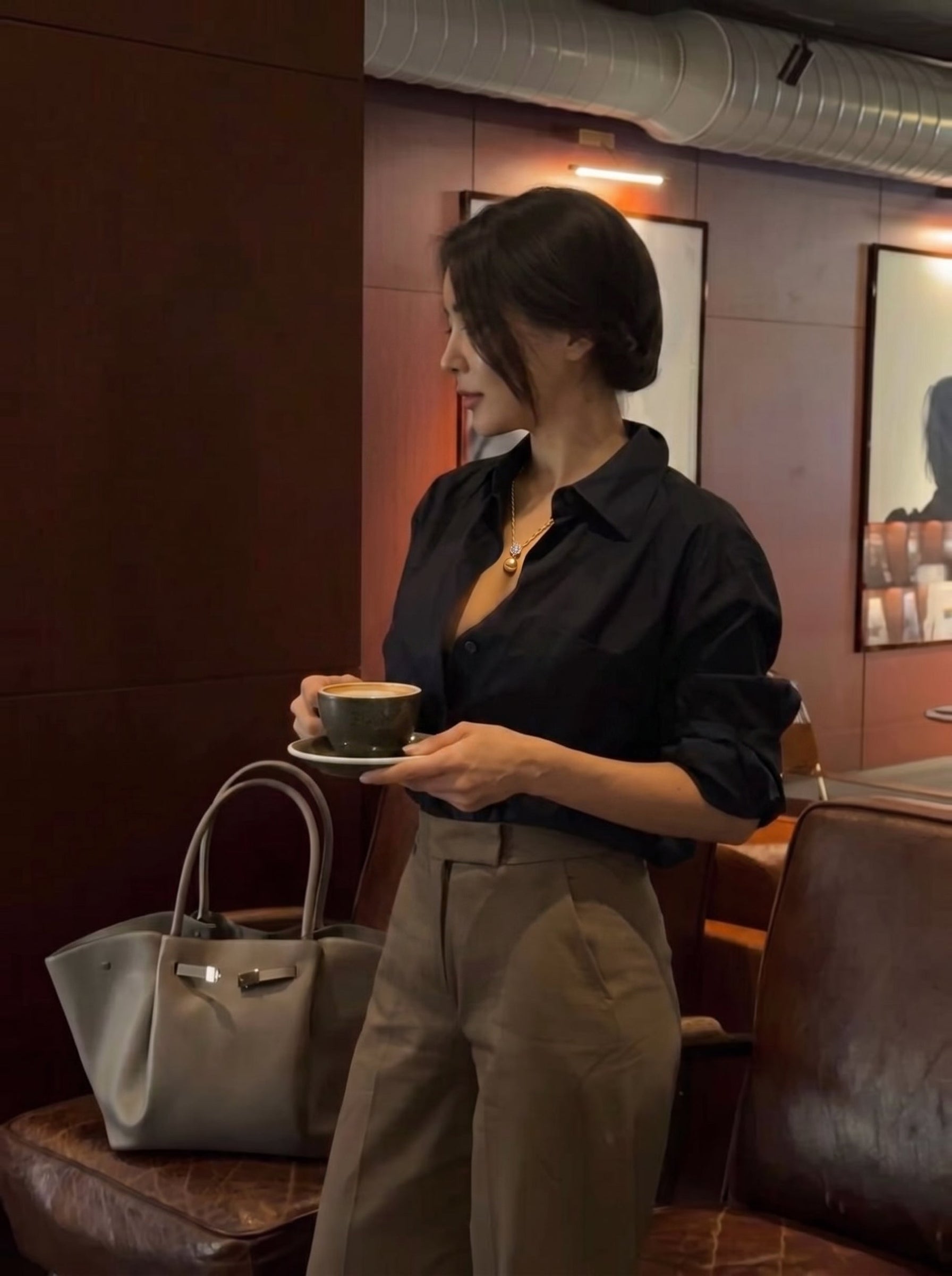 Une femme en chemise noire et pantalon marron tient une tasse de café tout en portant le collier pendentif unique carré Aurelia en perle des mers du Sud, présentant une grande perle dorée suspendue à une bélière carrée avec des pierres accent. Un élégant sac tote gris est posé à côté d'elle. @lifestyle
