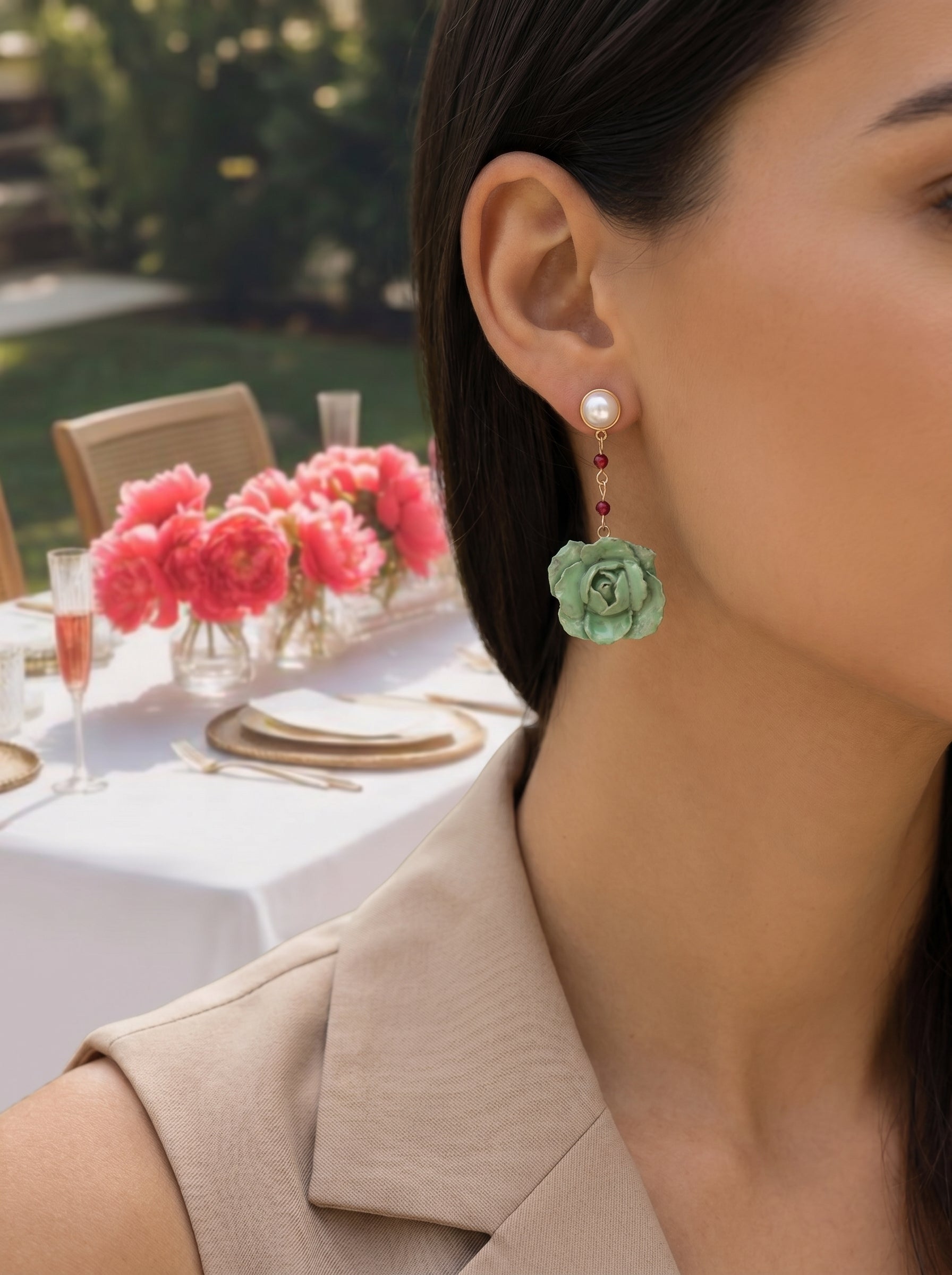 A model wears Awaken Freshwater Pearl Flower Dangle Earrings featuring a white pearl stud, a delicate chain with red beads, and a mint green flower pendant, with a blurred background of a garden party table setting. @lifestyle #lifestyle