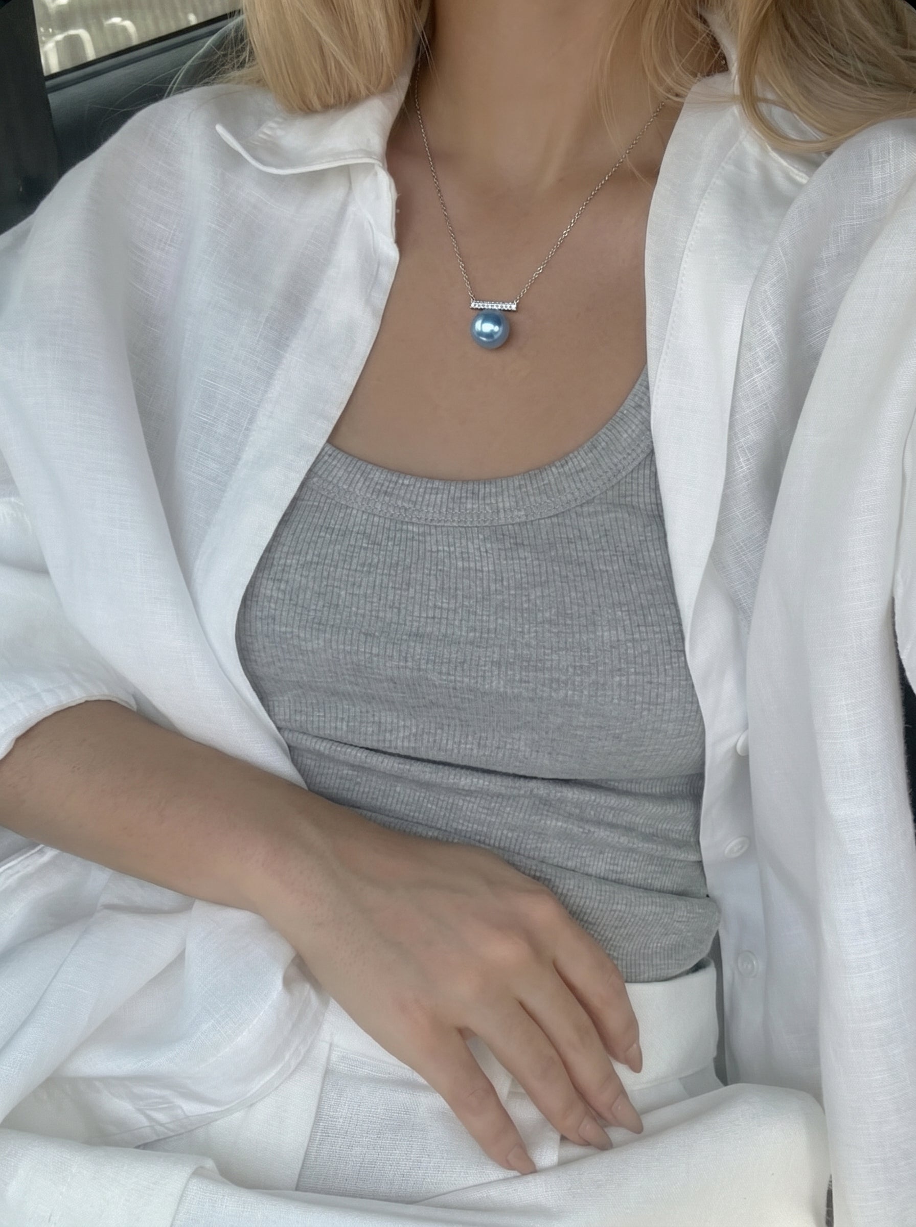 Une femme portant un débardeur gris et une chemise blanche ouverte, révélant le collier pendentif barre Azure en perle Akoya avec une seule et lustrée perle Akoya bleue suspendue à une délicate barre accentuée de pierres transparentes. @lifestyle