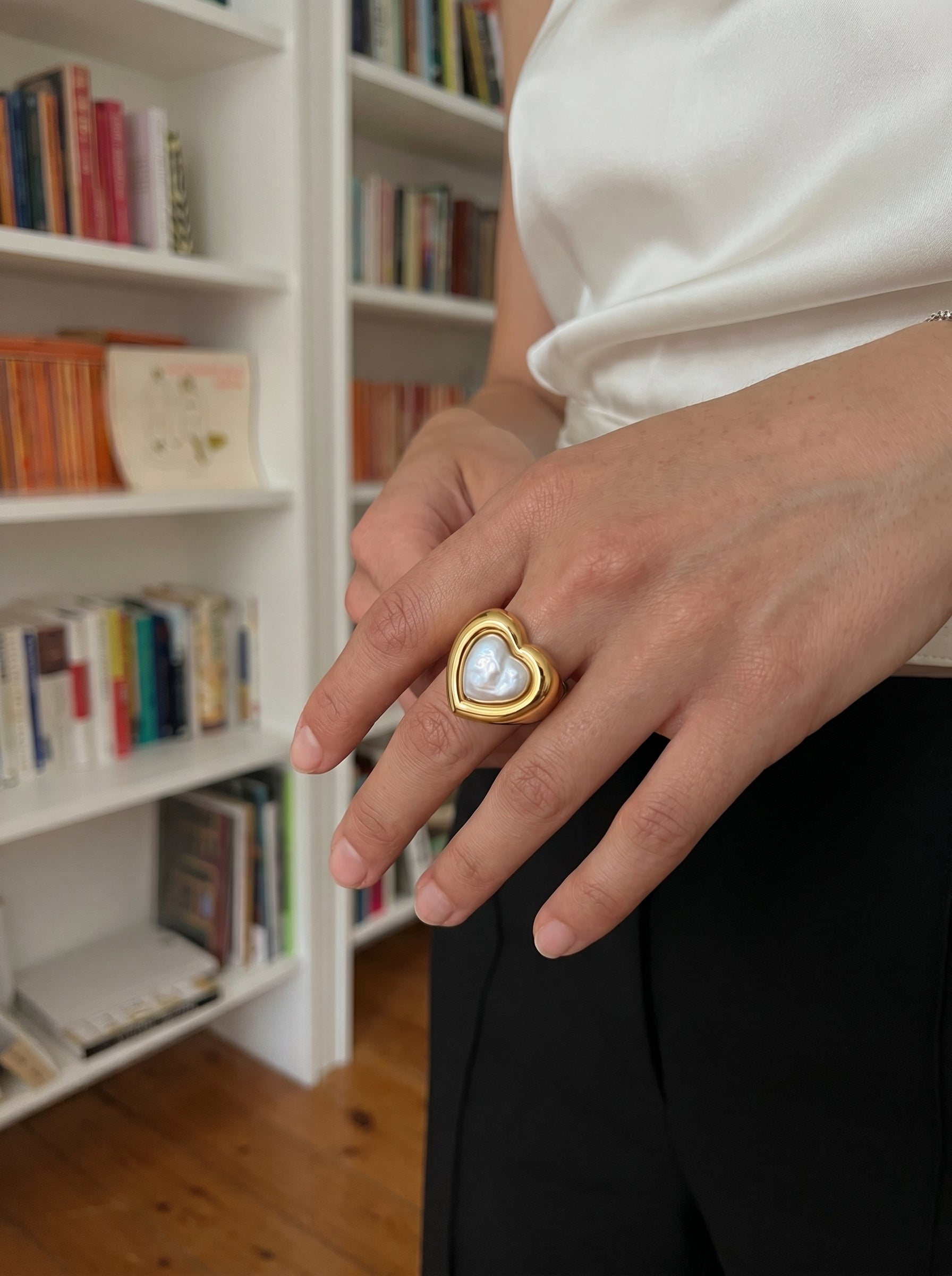A woman's hand wears a bold gold cuff ring featuring a heart-shaped bezel with a white freshwater pearl, set against a backdrop of bookshelves. @lifestyle #lifestyle