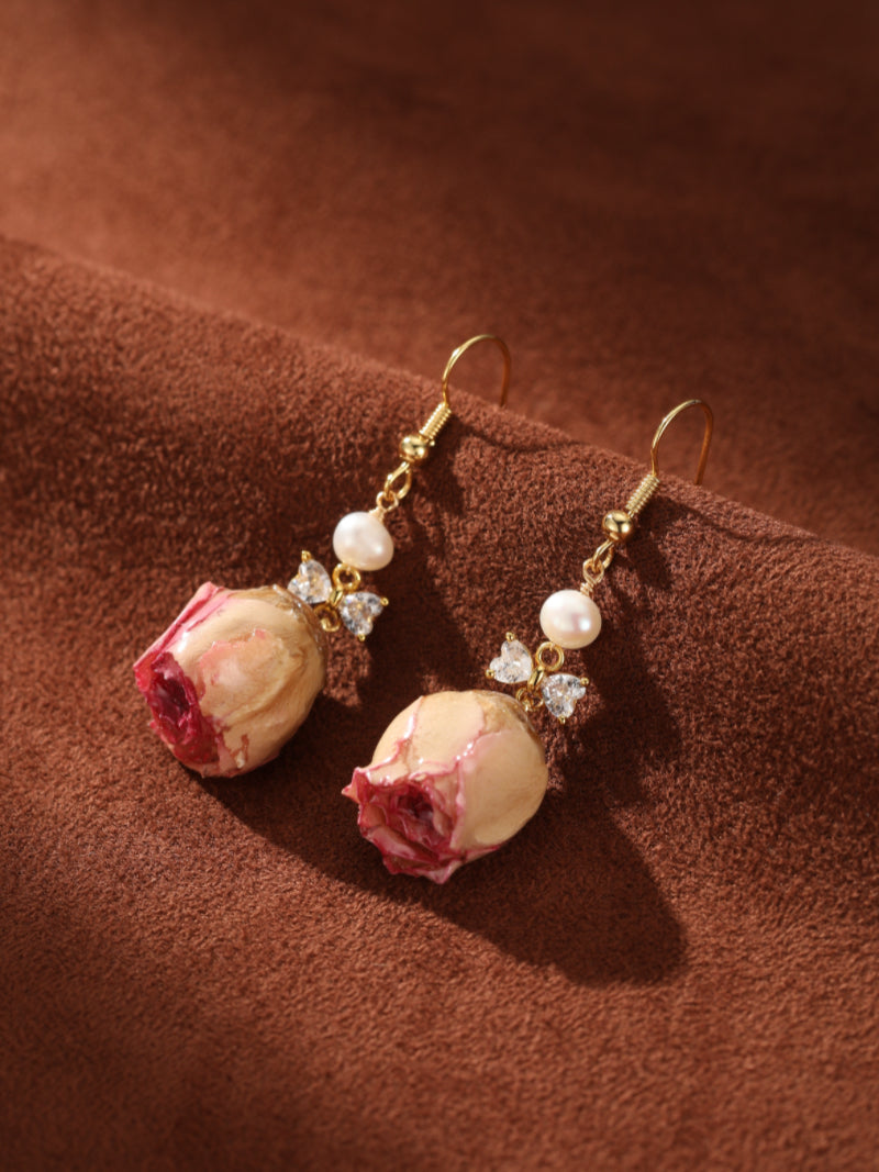 Botanical Dew Freshwater Pearl Flower Dangle Earrings feature a dried pink rosebud, a lustrous white pearl, and clear bow-shaped accents, all suspended from gold-toned hooks. @pearl