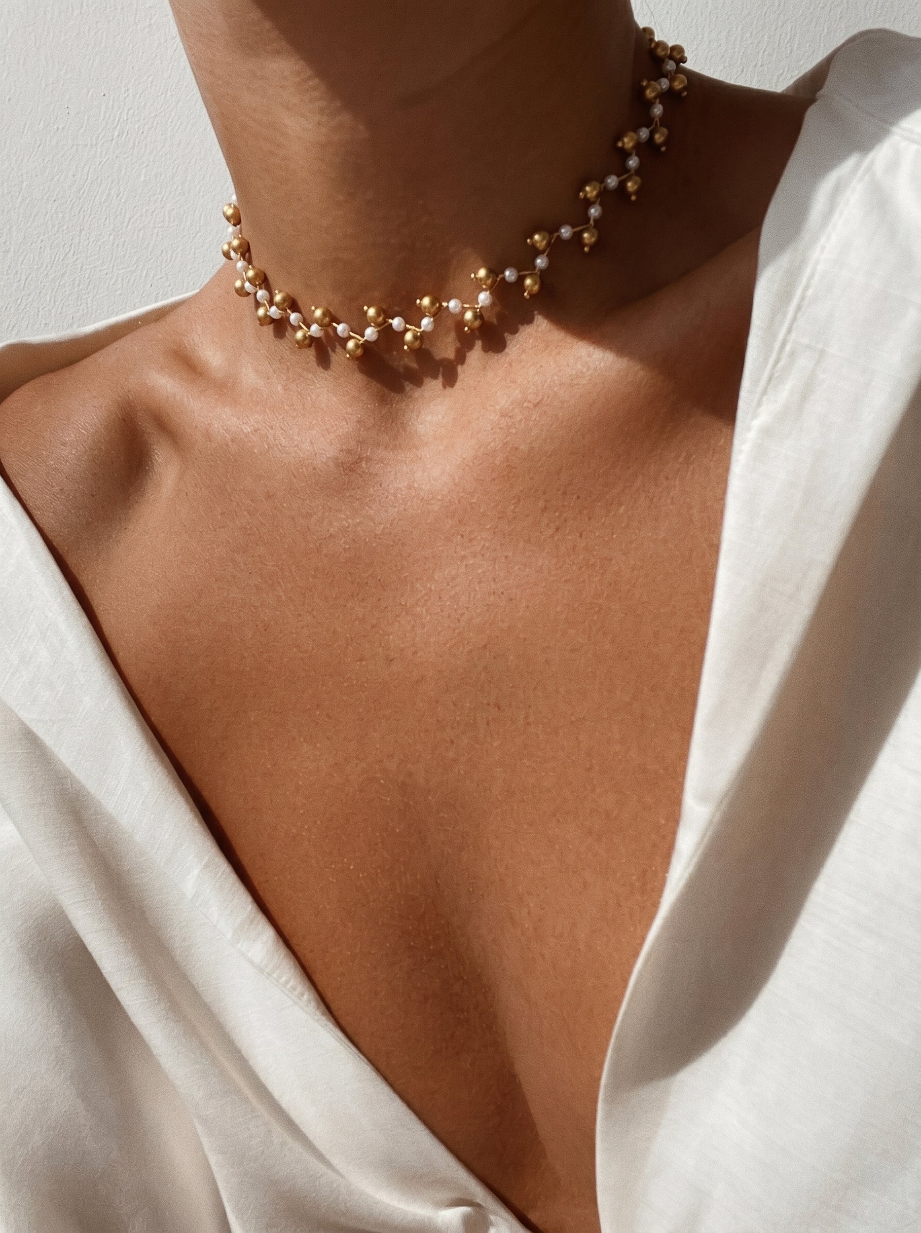 A close-up shot of a model wearing the Branch Weave Mother of Pearl Twist Beaded Necklace with delicate gold accents and small white beads, layered on a white shirt. @model