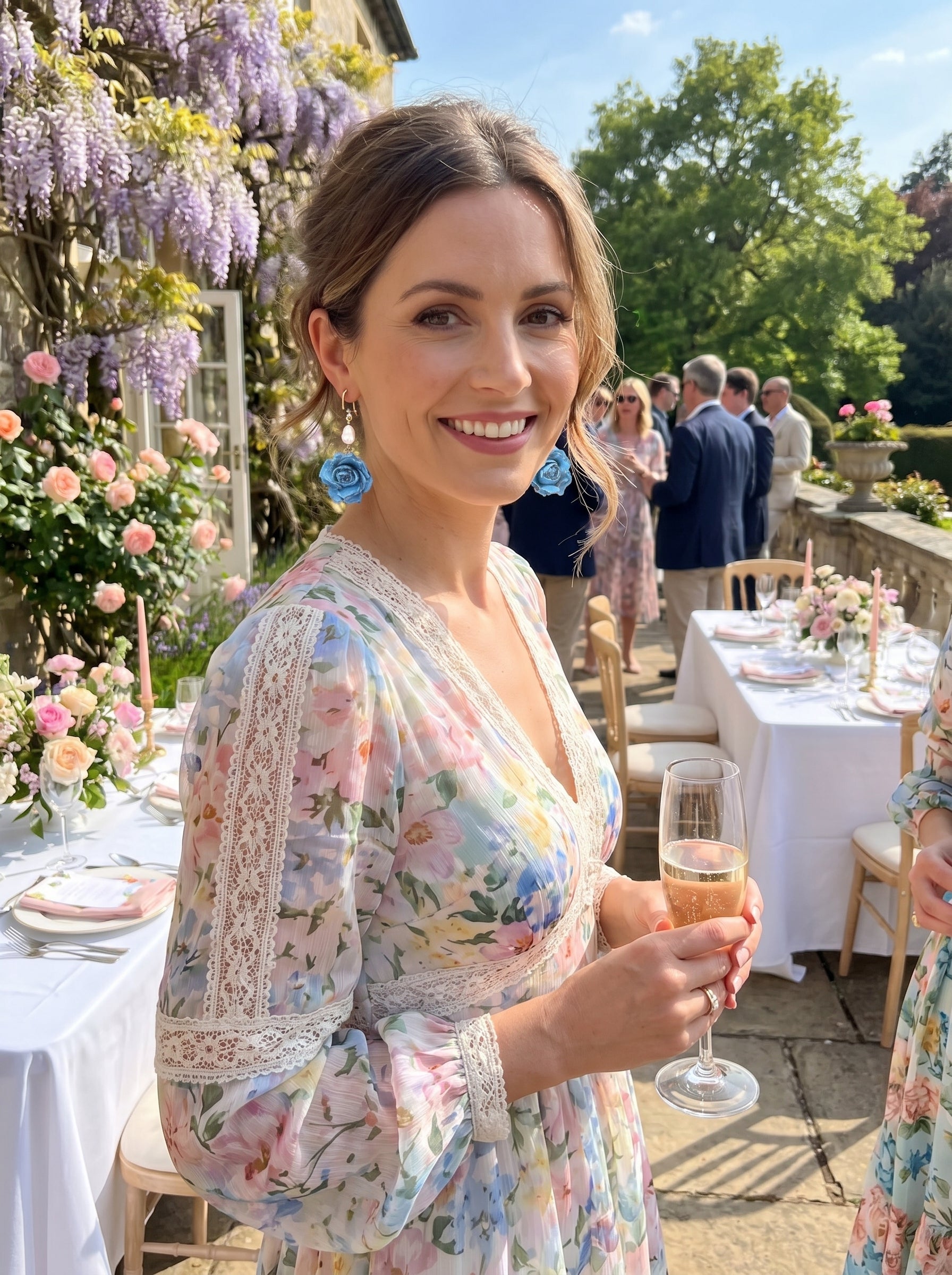 A woman smiles while wearing Celeste Reverie blue flower and pearl dangle earrings, a floral dress, and holding a champagne flute at an outdoor event with wisteria and tables set for a meal. @lifestyle #lifestyle