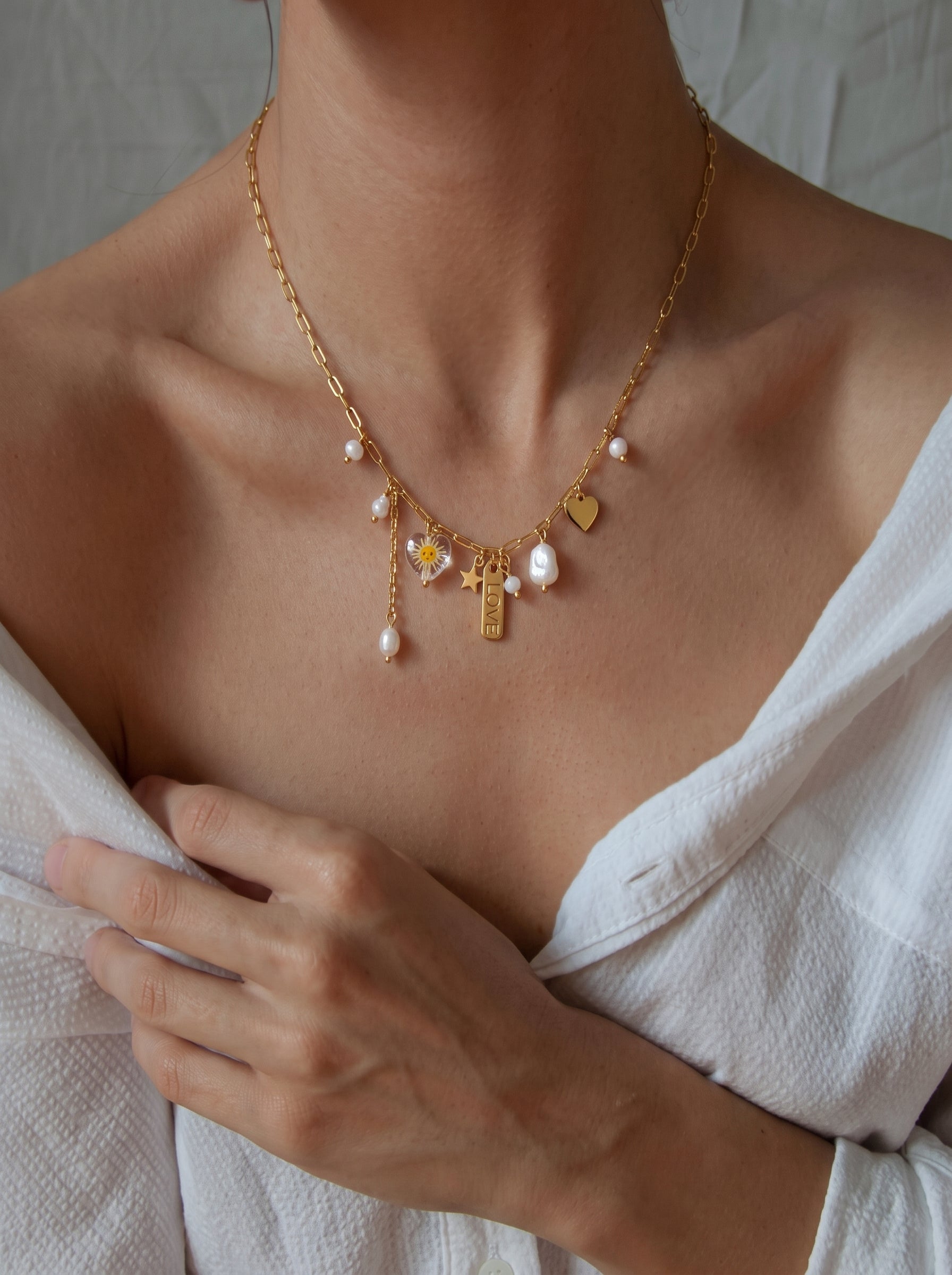 A gold-tone necklace with a paperclip chain, featuring floating freshwater baroque pearls, a small gold star, a heart charm, and a rectangular tag engraved with the word LOVE. A daisy charm is also visible. The necklace is worn by a model and is part of a Celestial Whim collection. @model