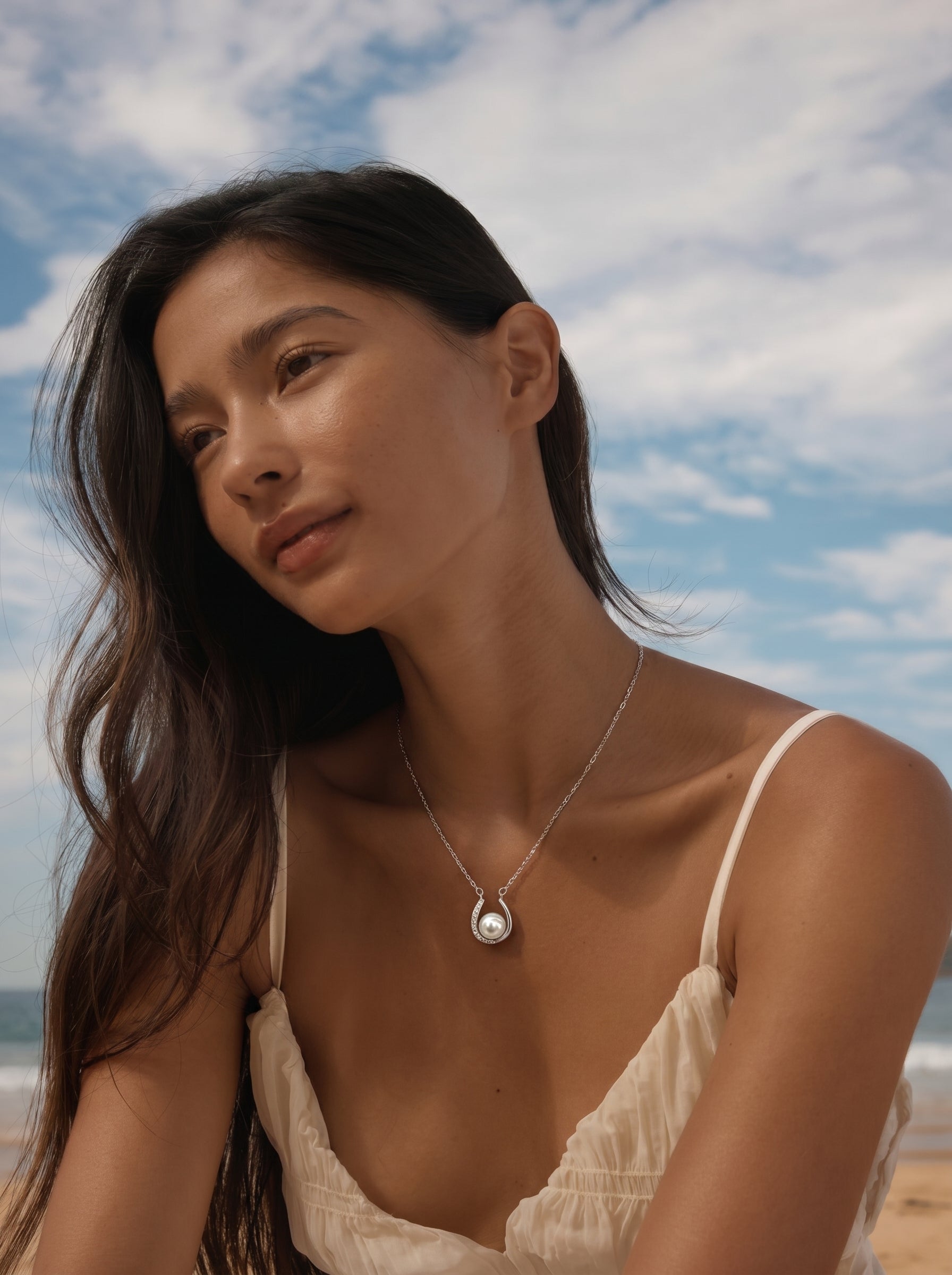 Une femme porte un collier à chaîne argentée avec un pendentif fer à cheval berçant une lumineuse perle Akoya, contre un fond de plage et de ciel bleu. @lifestyle