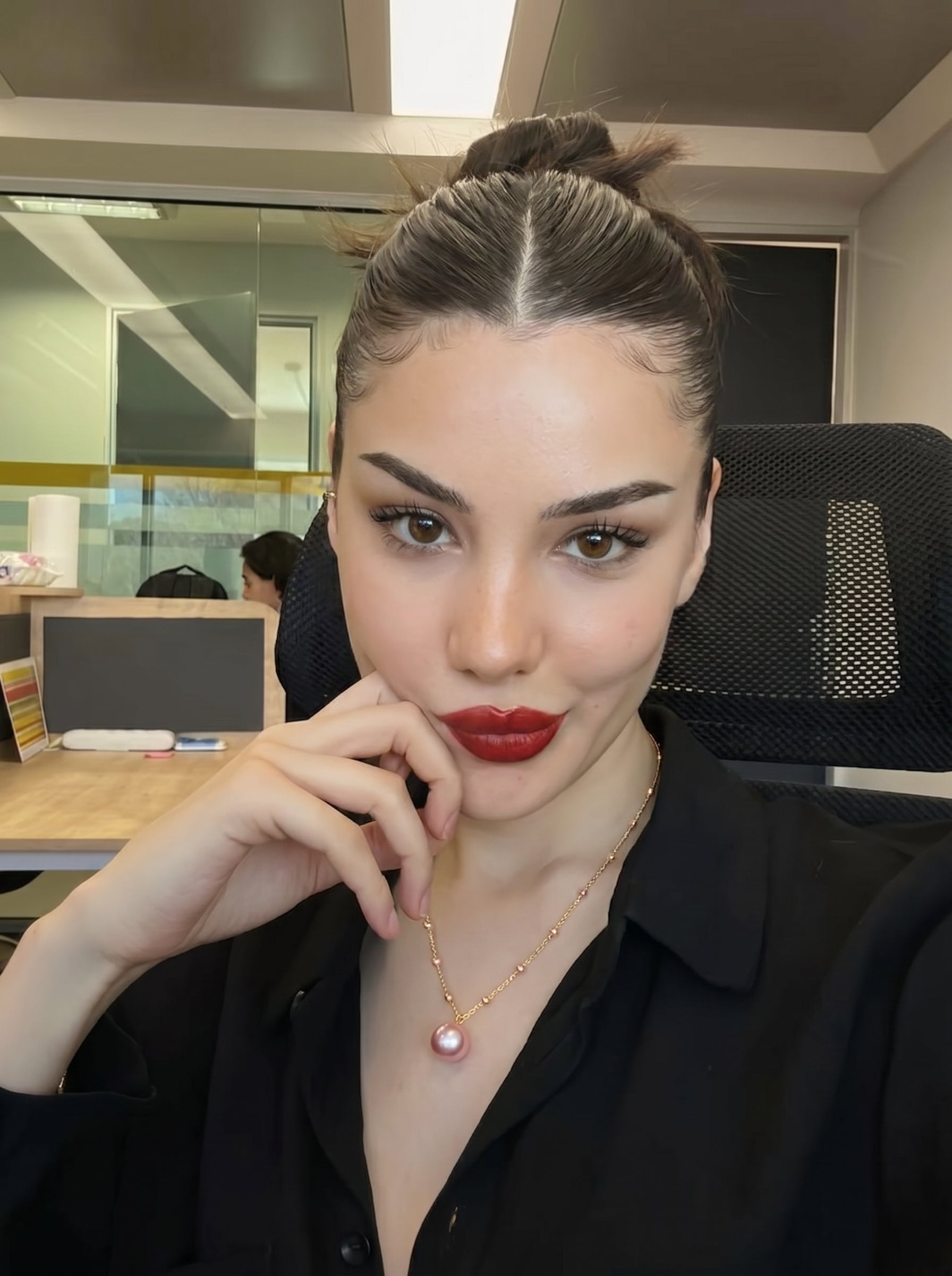 A woman wearing a black shirt and a delicate gold-tone necklace with a large pink freshwater pearl pendant and smaller floating pearls. She is in an office setting, looking at the camera with her hand near her chin. @model #lifestyle