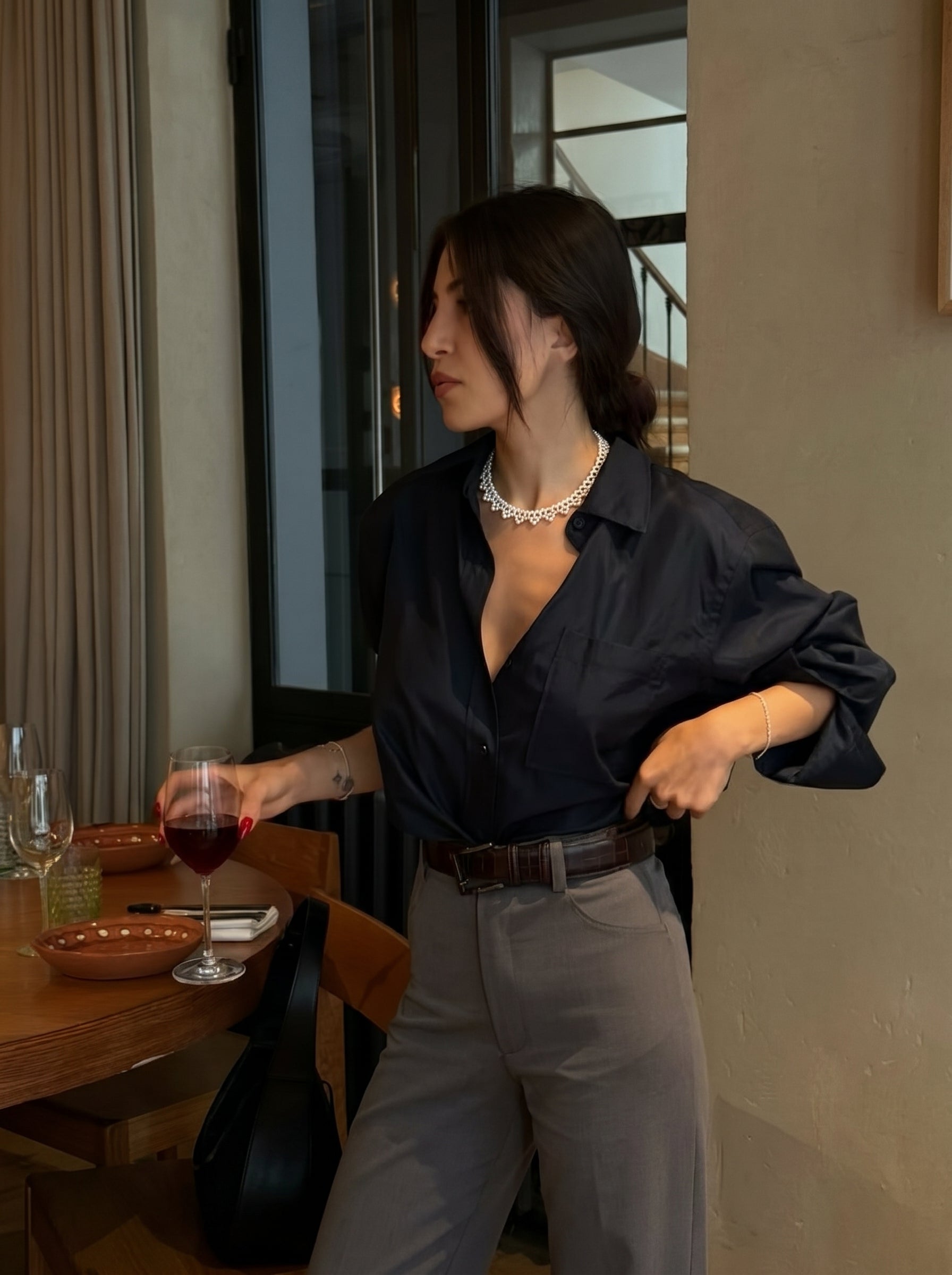 Une femme portant une chemise noire, un pantalon gris, et le collier ras-de-cou tissé Cipher en perle d'eau douce, tenant un verre de vin rouge à une table. @lifestyle