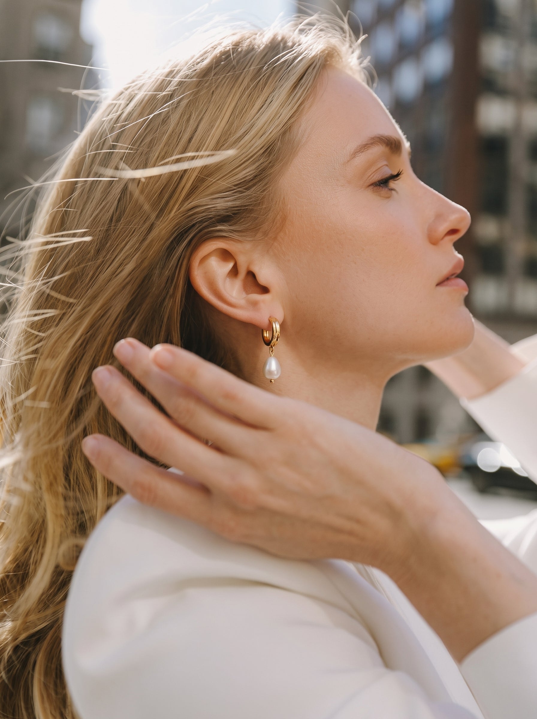 A blonde woman wearing a white blazer and gold hoop earrings with a single white freshwater pearl drop is shown from the side in an urban setting. @lifestyle