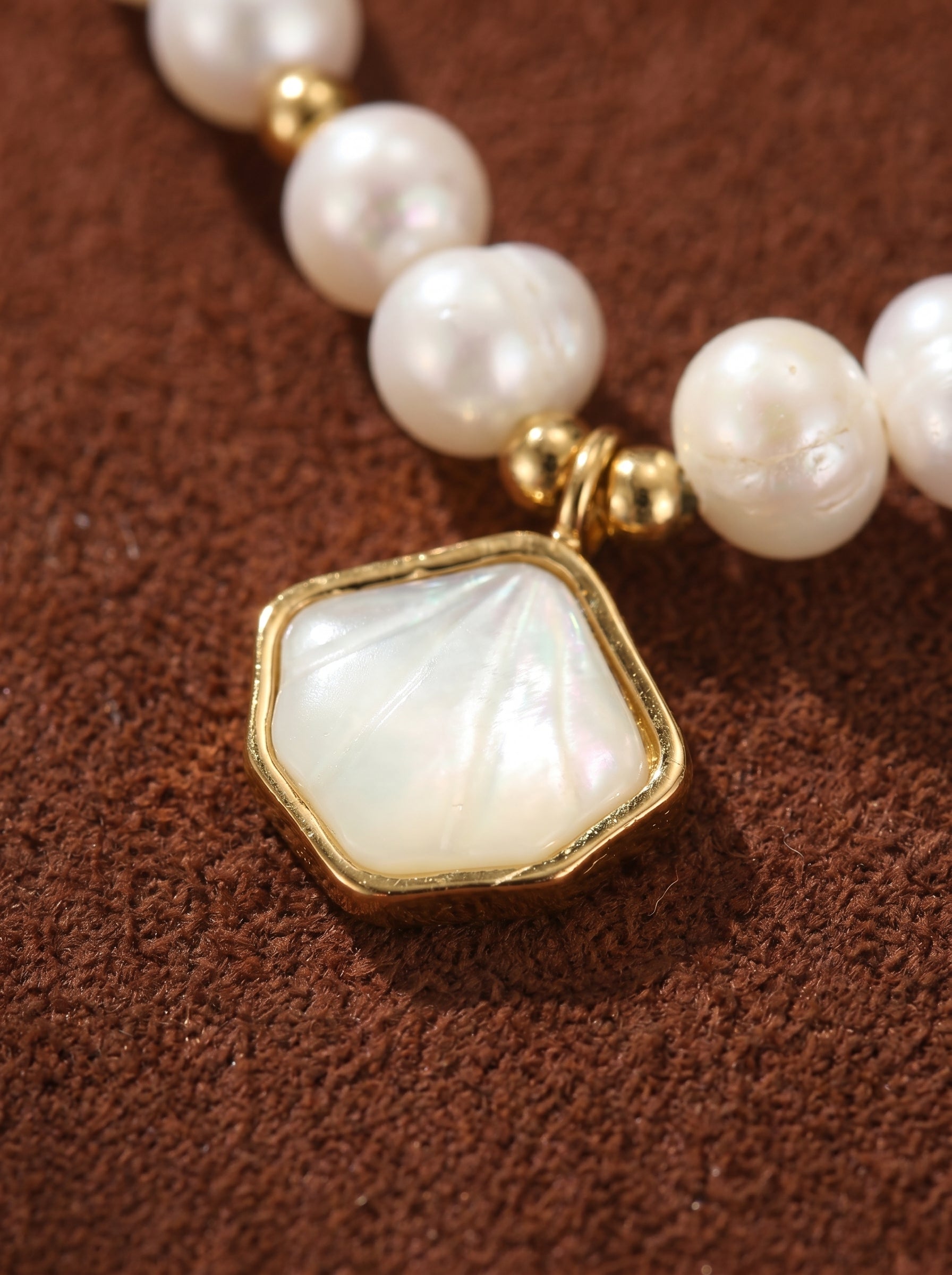Close-up of a bracelet featuring smooth white freshwater pearls and a gold-trimmed, iridescent seashell pendant on a textured brown background.@pearl