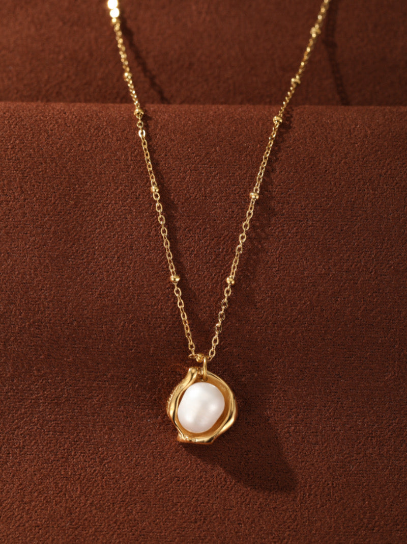 Collier pendentif Cocoon présentant une perle blanche d'eau douce sertie dans un vase sculptural ton or sur une délicate chaîne perlée ton or.