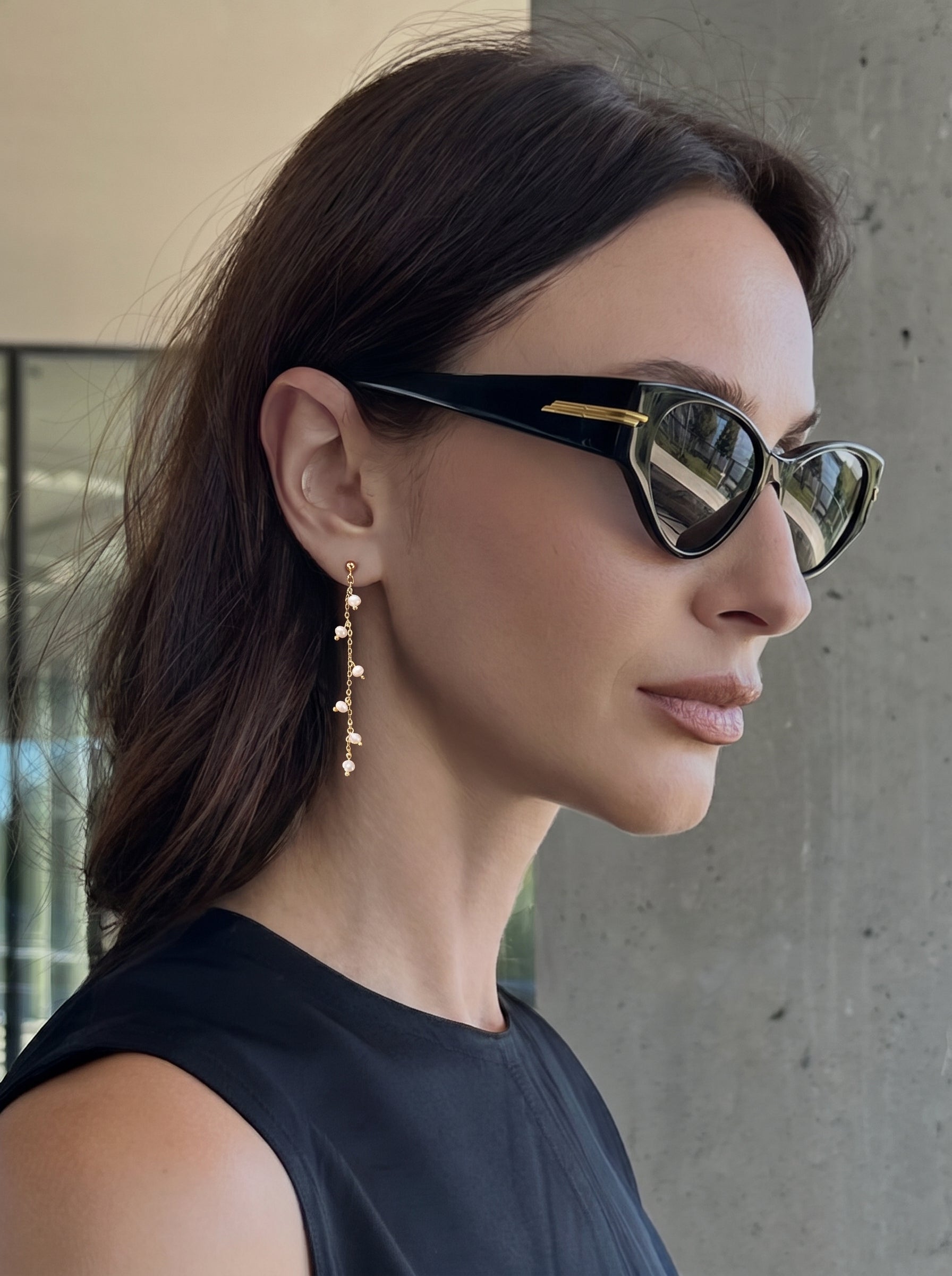 Close-up of a woman wearing stylish black sunglasses and a black top, showcasing delicate gold chain drop earrings adorned with small, luminous freshwater pearls. @lifestyle #lifestyle