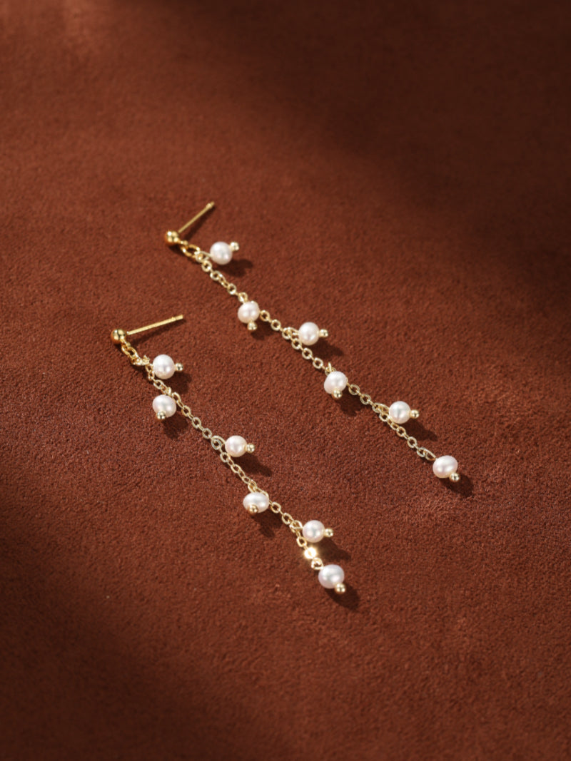 Gold chain drop earrings with multiple small, seed-shaped freshwater pearls cascading down, set against a brown background. @pearl
