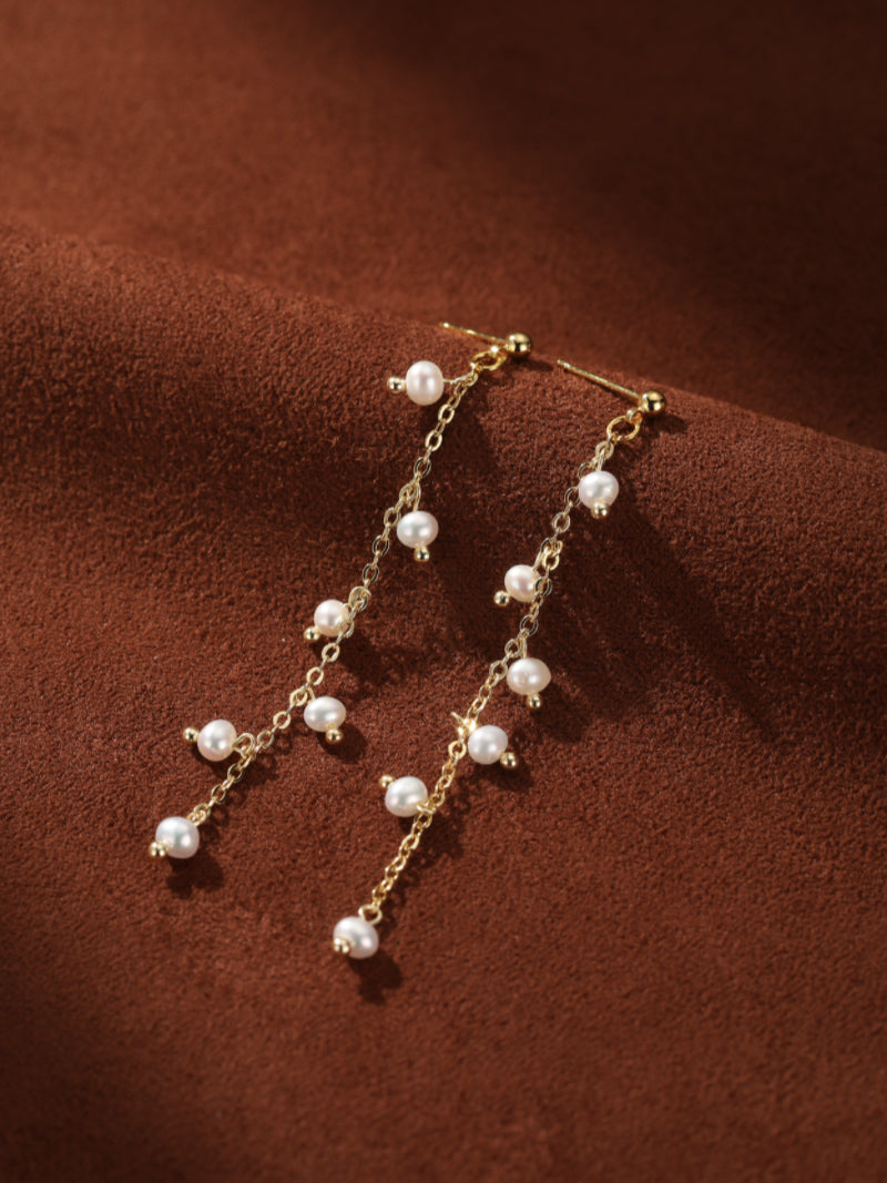 Delicate gold chain drop earrings featuring multiple small, white freshwater seed pearls cascading down the chains against a textured brown background.