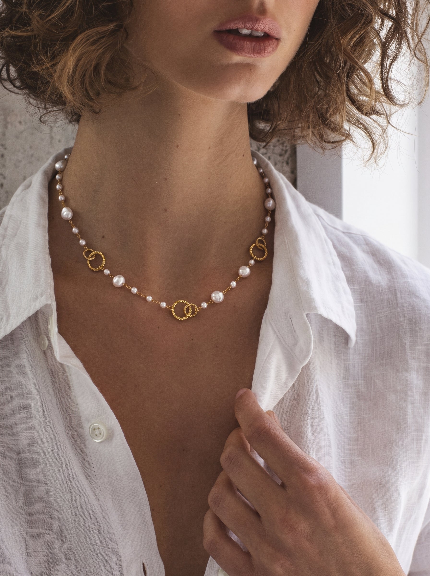 A woman wearing a white linen shirt and the Constellation Imitation Pearl Ring Long Floating Necklace, featuring a gold-tone chain with varying sizes of white imitation baroque pearls and textured gold-tone rings. @model