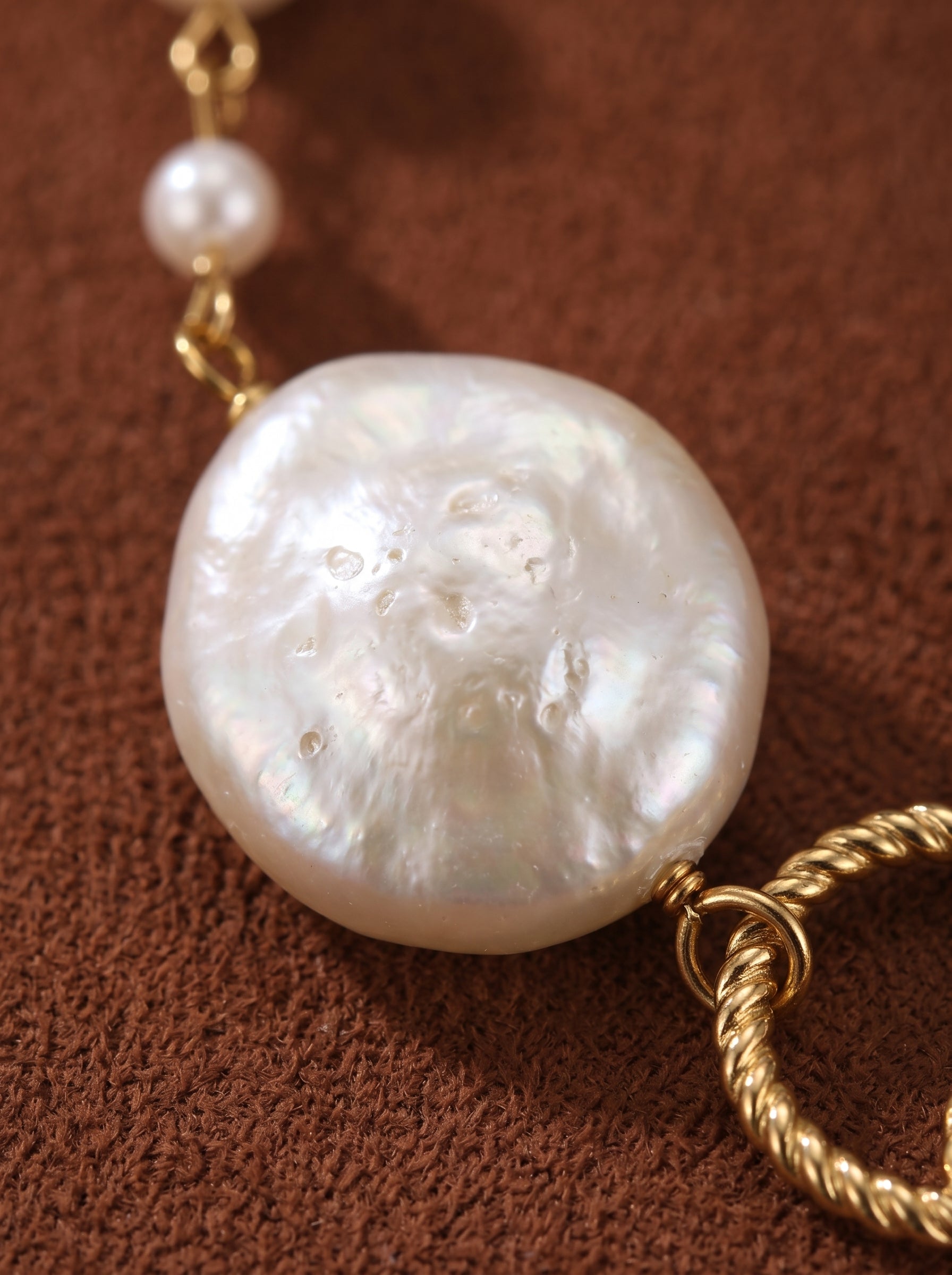 Close-up of a Constellation imitation pearl ring long floating necklace with a large, irregular white baroque pearl and textured gold rings on a brown textured background. @pearl
