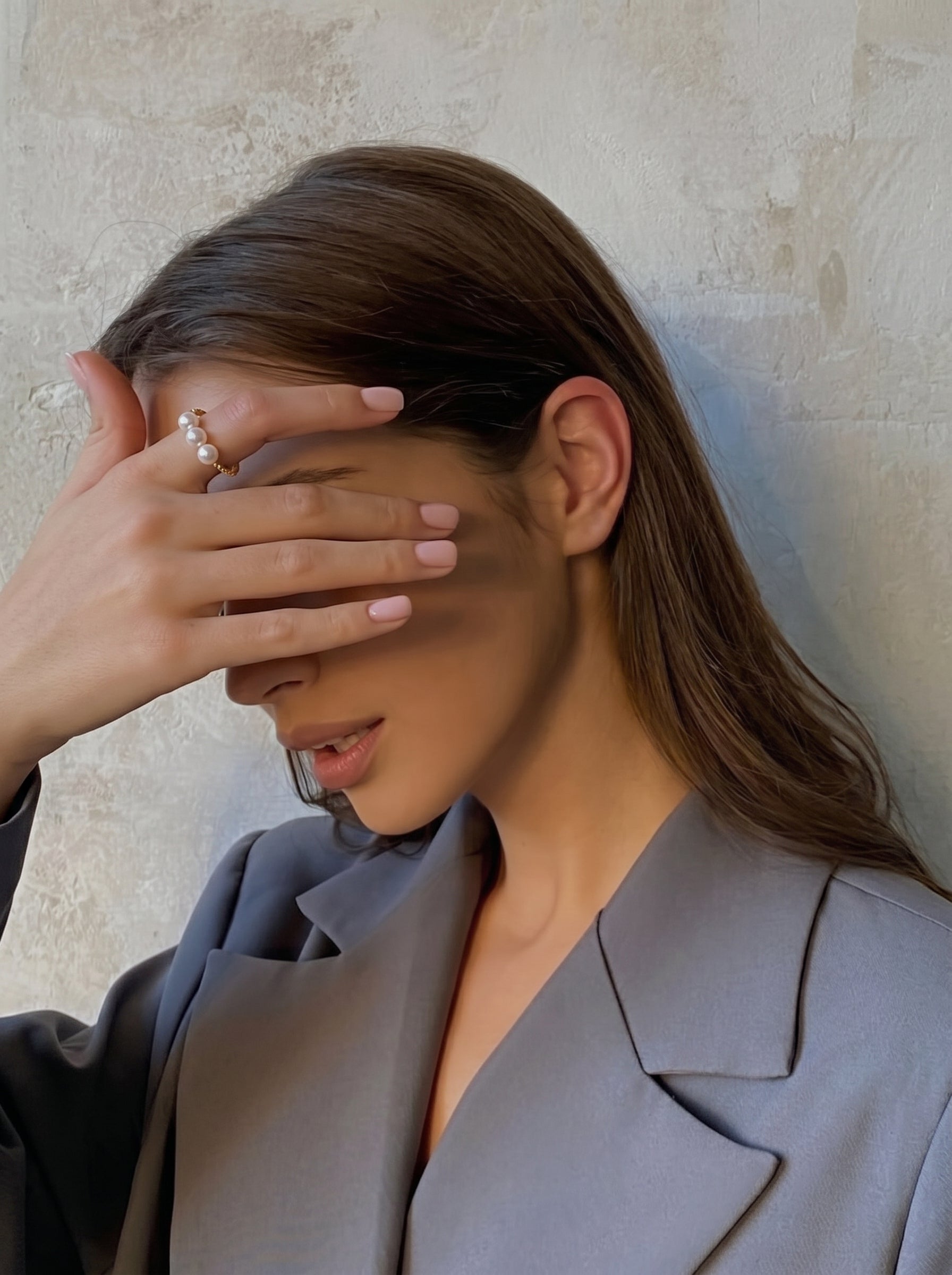 A woman's hand, adorned with the Continuum Freshwater Pearl Cuff Ring, is held up to her face, partially obscuring her eyes. The ring features a twisted band and three small, round white freshwater pearls. She is wearing a grey blazer. @model