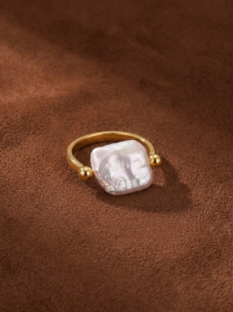 Gold-toned open cuff ring featuring a large, irregular white freshwater pearl with a textured band and two small gold balls on either side of the pearl. The ring is displayed on a textured brown surface with soft shadows.