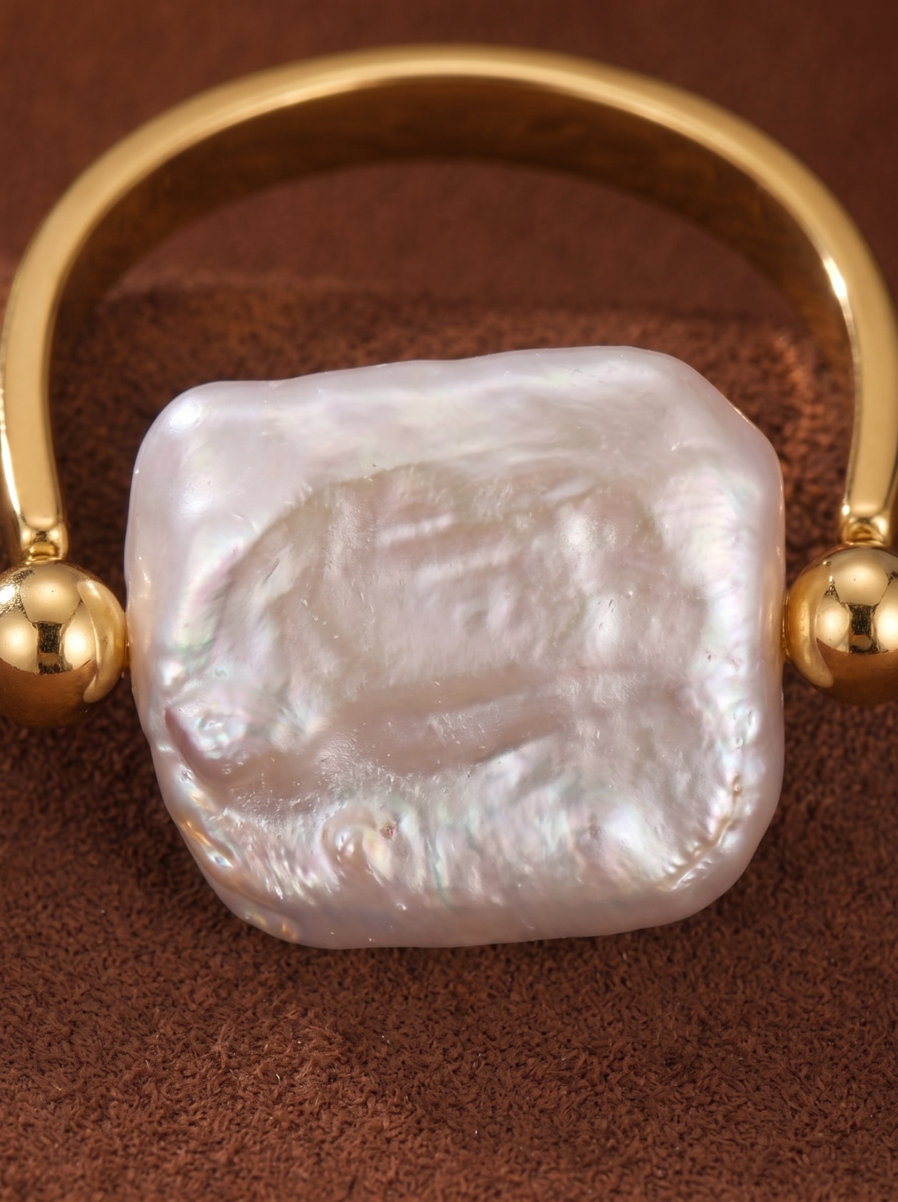 Gold-toned open cuff ring featuring a large, irregular white freshwater pearl with a lustrous sheen, accented by two small gold beads on a brown textured background. @pearl