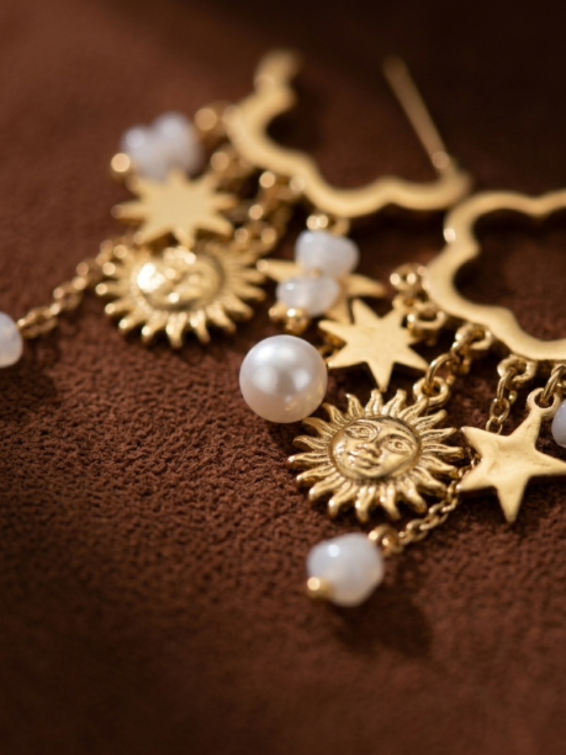Gold dangle earrings featuring freshwater pearls, sun charms with faces, and star charms against a brown textured background.