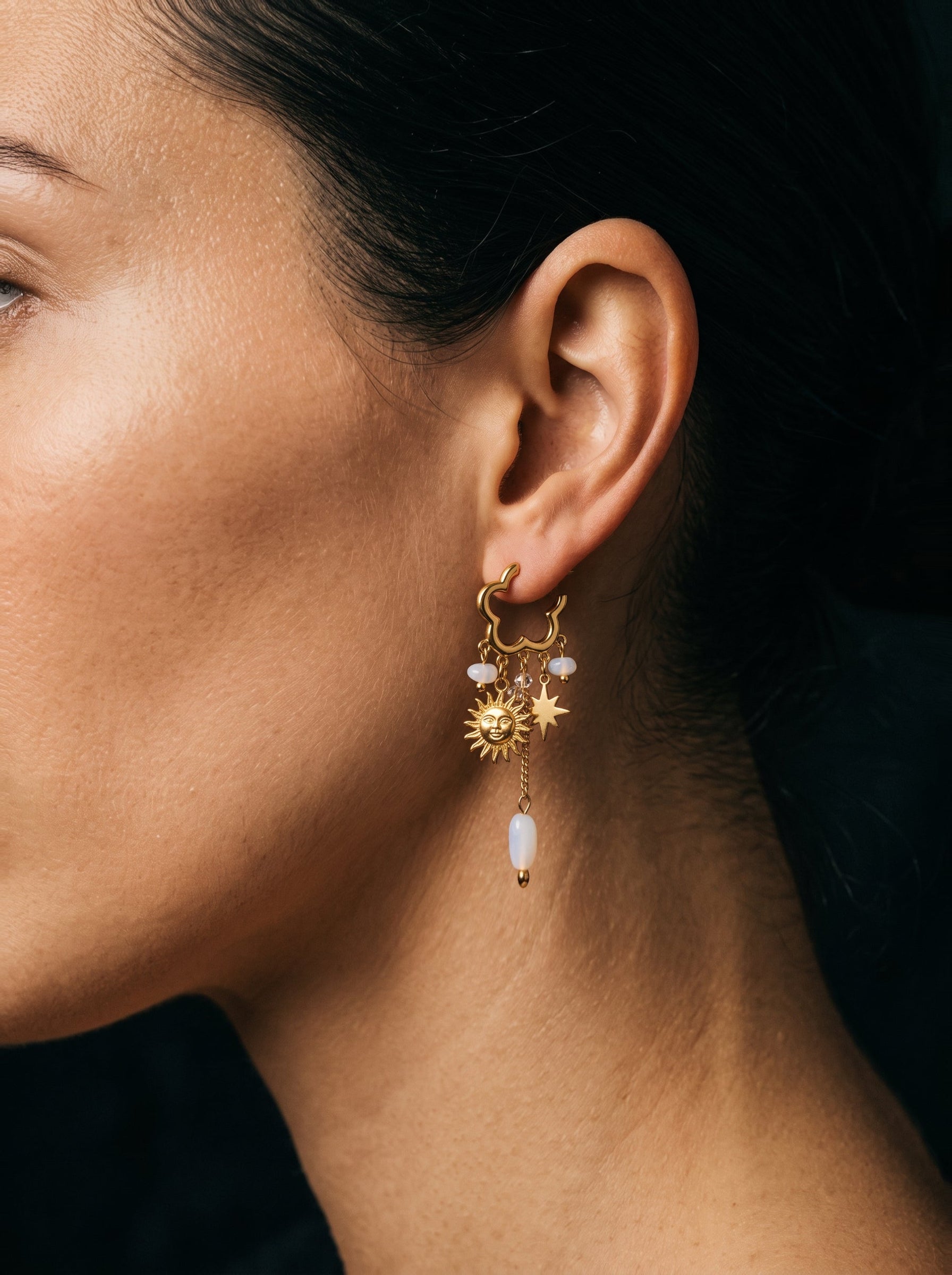 Gros plan d'une mannequin portant des boucles d'oreilles pendantes dorées présentant des perles d'eau douce, une breloque soleil, et une breloque étoile. @model