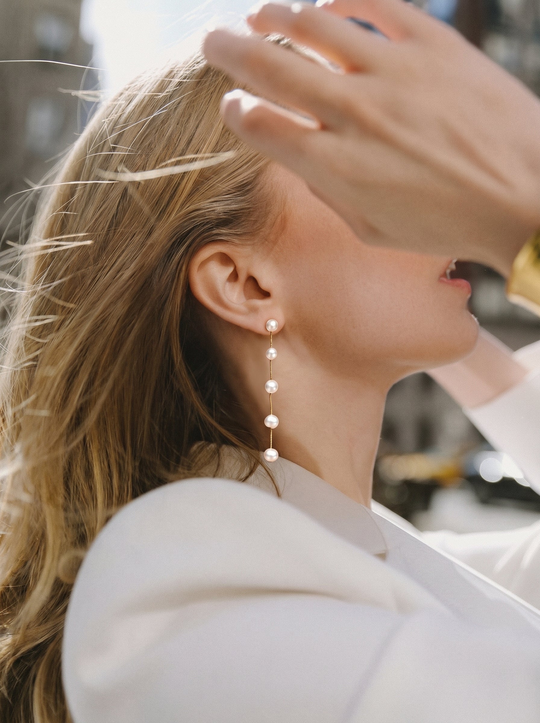 A woman with blonde hair is wearing delicate gold-toned dangle earrings featuring five spaced white freshwater pearls. The earrings are a linear drop design, and the shot is a close-up with a soft, blurred urban background. @lifestyle