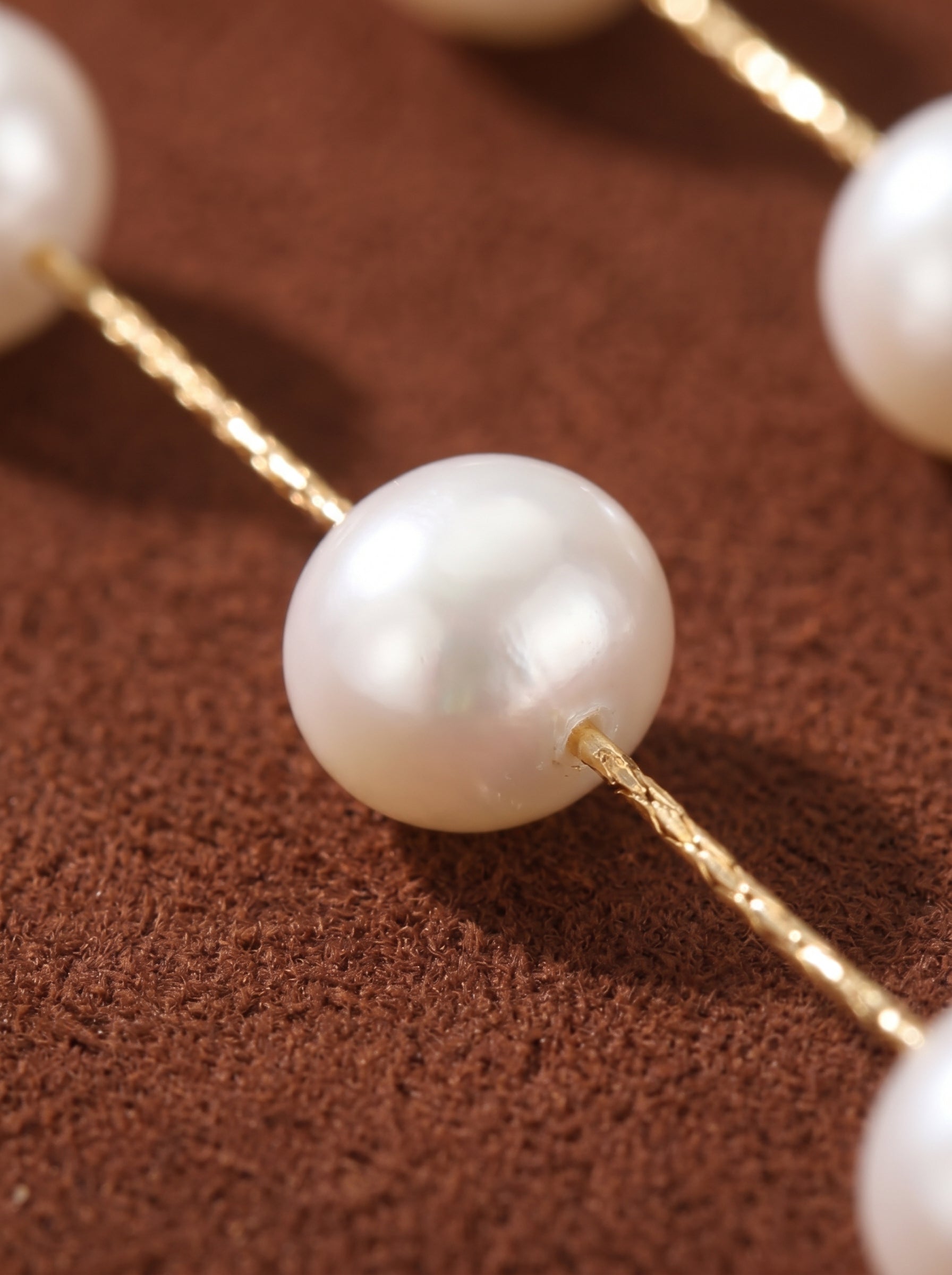 Close-up of white freshwater pearl dangle earrings featuring lustrous 6mm button pearls spaced along delicate gold-toned strands on a brown textured background. @pearl