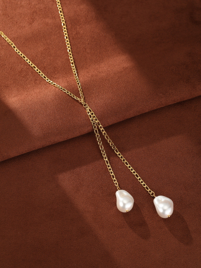 Gold-toned chain lariat necklace with two irregular white imitation Baroque pearls hanging at the ends, displayed on a brown textured background.