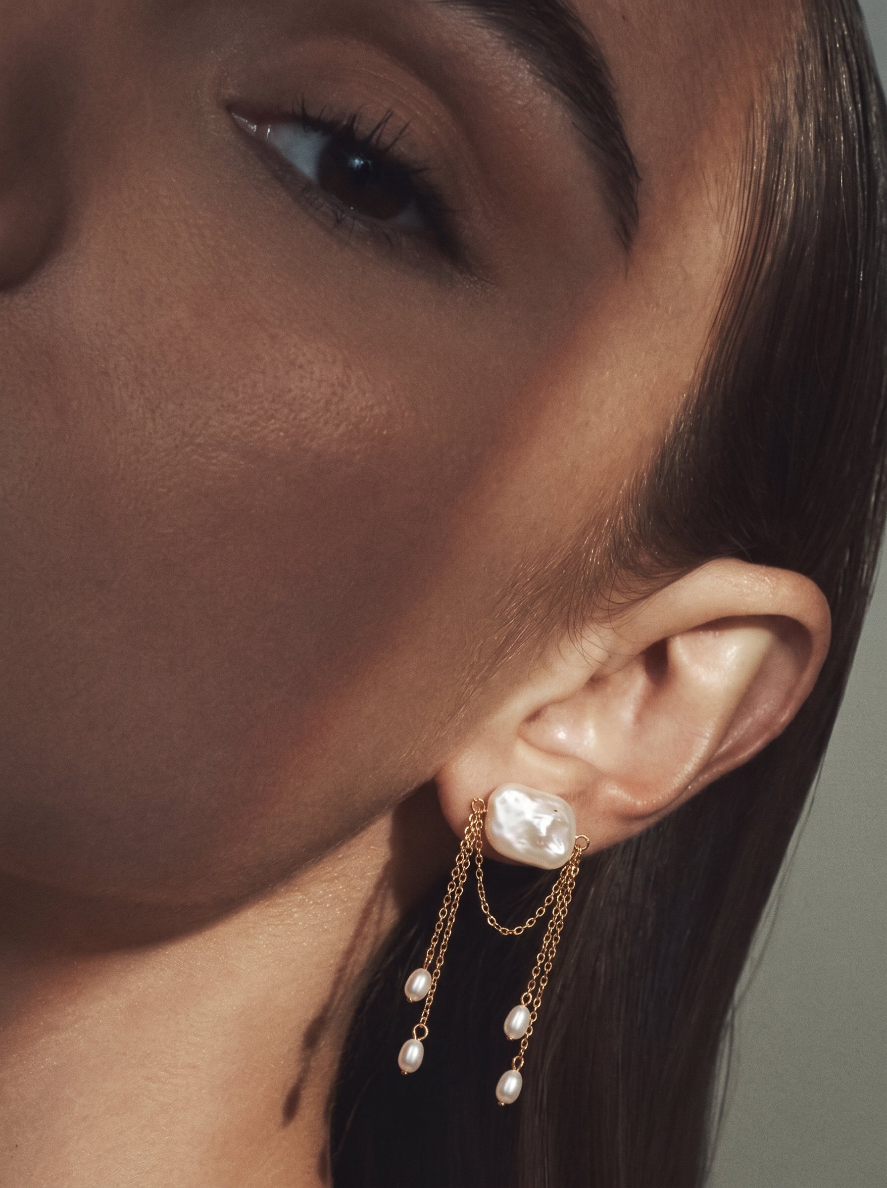 Close up of a model wearing Driftfall Freshwater Pearl Fringe Drop Earrings, featuring a large baroque pearl connected by delicate gold chains to smaller pearl drops. @model