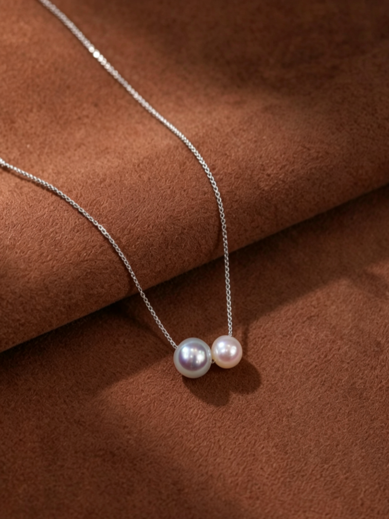A delicate silver chain necklace featuring two Akoya pearls, one 7mm and one 9mm, appearing to float closely together against a textured brown background.