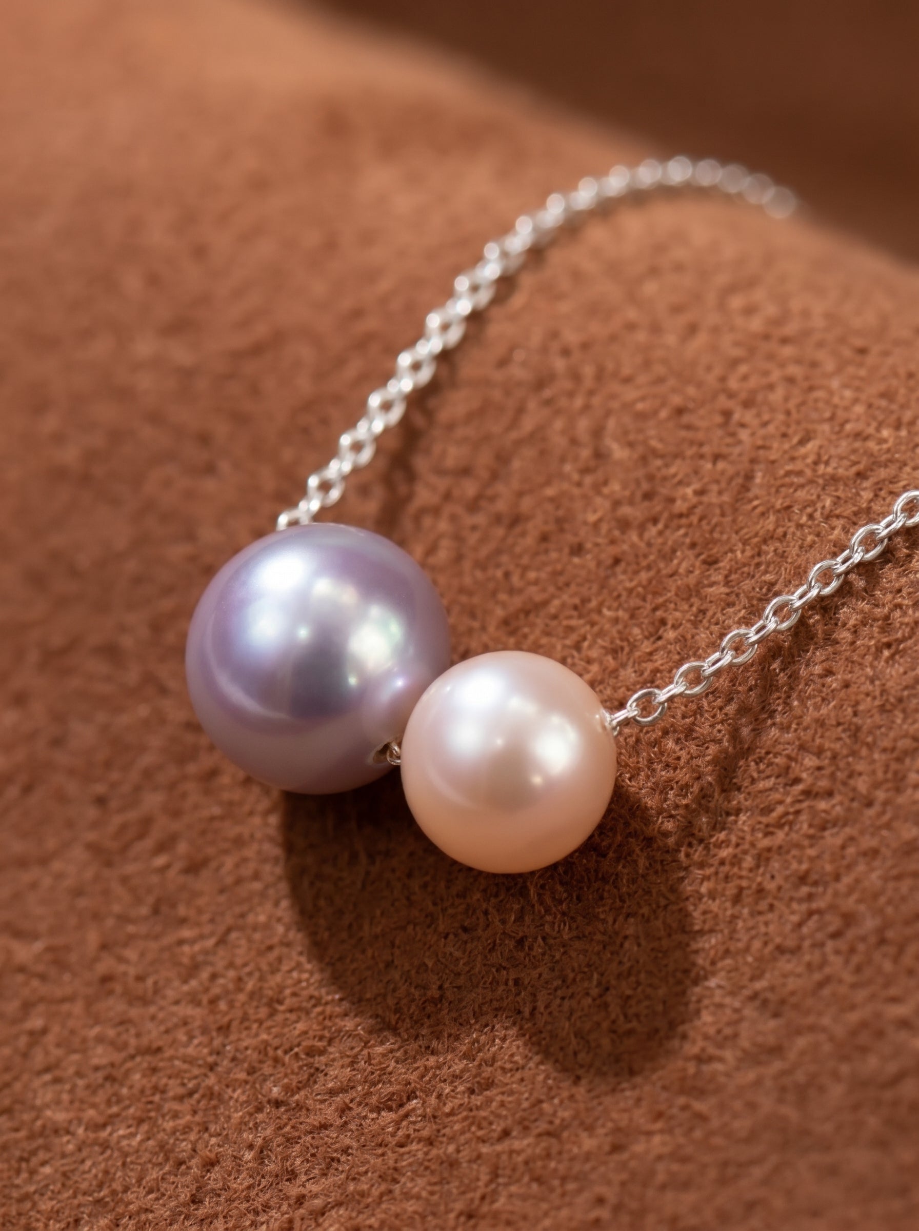 A delicate silver chain necklace featuring two Akoya pearls, one 7mm lavender pearl and one 9mm peachy-pink pearl, appearing to float. The pearls have iridescent overtones. @pearl