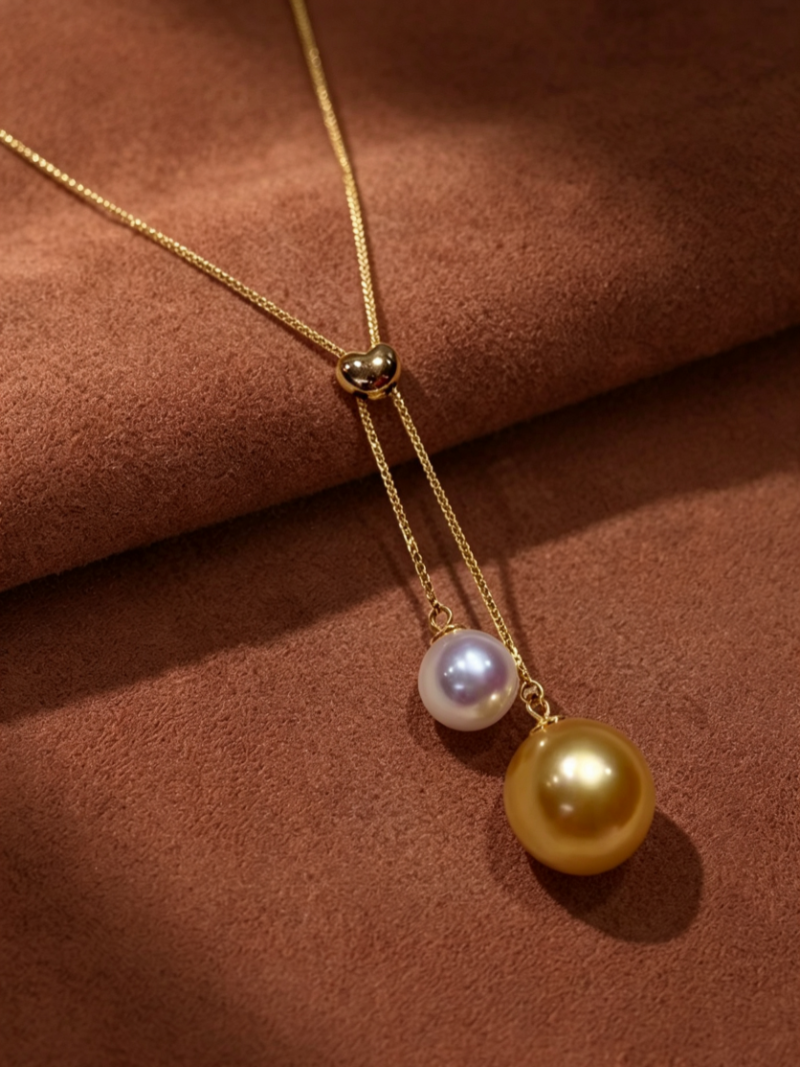 Duet necklace featuring a heart-shaped gold clasp and two pearls, one white Akoya and one golden South Sea pearl, on a fine gold chain against a brown textured background.