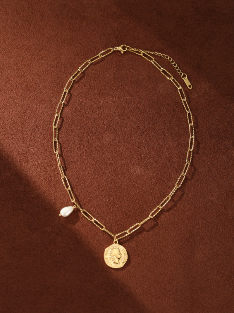 Collier à chaîne trombone imposante dorée présentant un pendentif pièce et une seule goutte de perle blanche d'eau douce sur fond texturé marron.