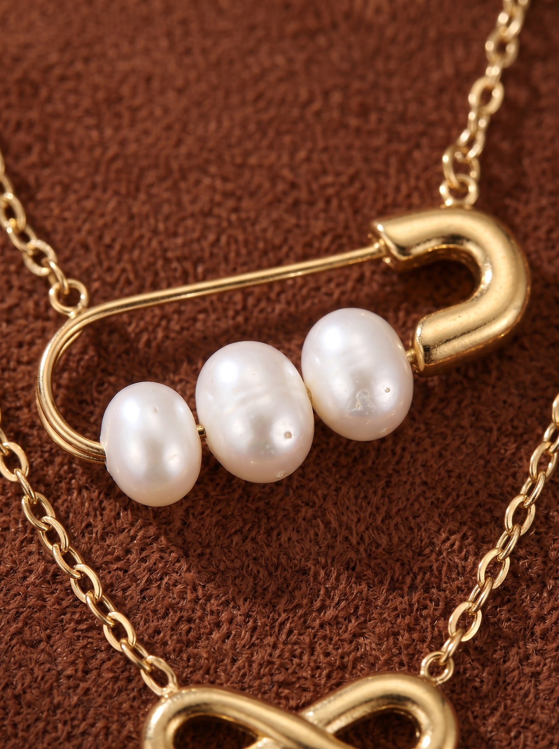 Collier superposé ton or présentant un symbole infini et une breloque épingle de sûreté tenant trois perles graines blanches d'eau douce sur fond texturé marron. @pearl