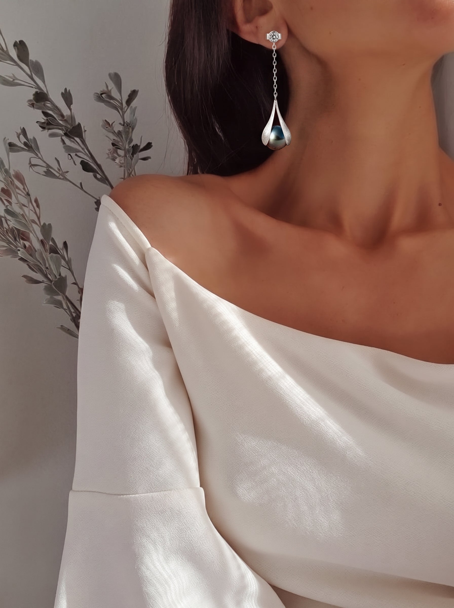 A woman models Enfold Tahitian Pearl Drop Earrings, featuring a 10mm round black pearl in a silver-tone setting hanging from a clear stone stud. The earrings are delicate and modern, complementing her elegant white off-the-shoulder top. @model
