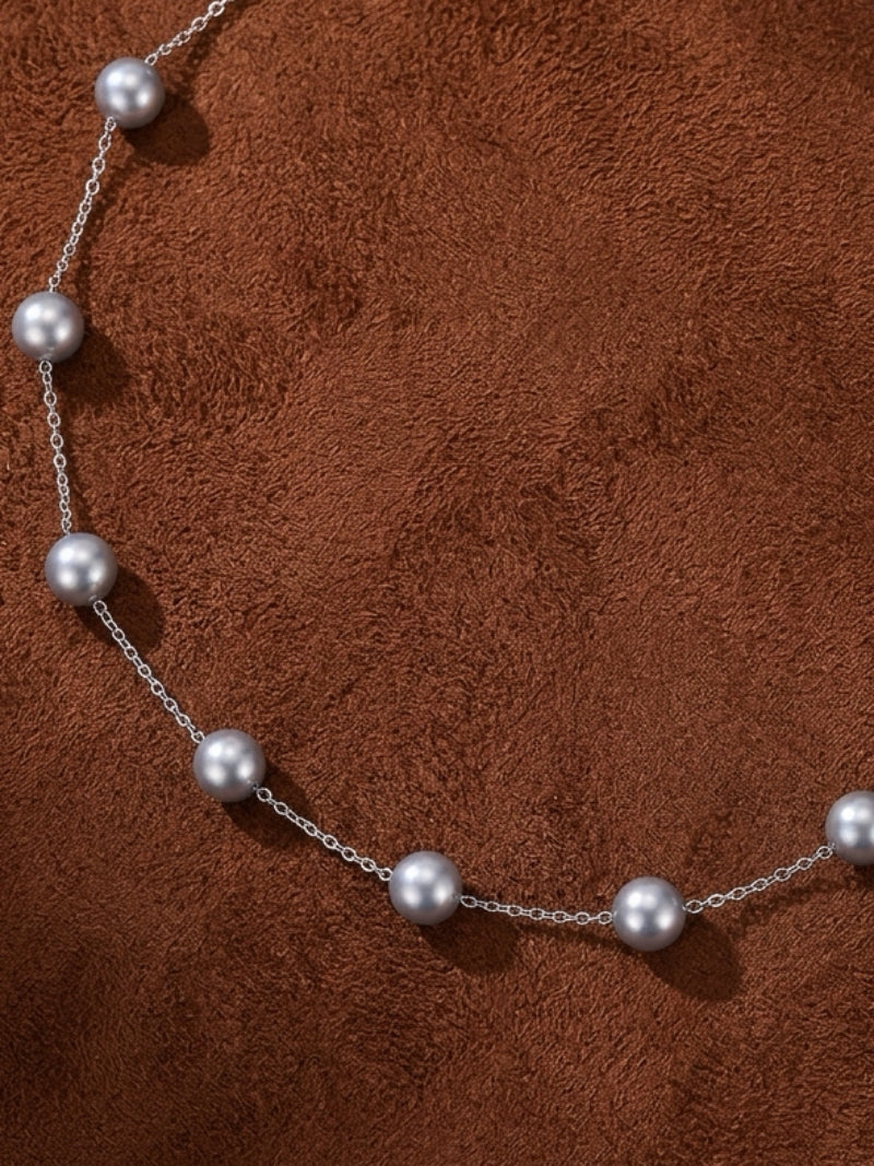 A delicate silver chain necklace with multiple spaced grey Akoya pearls against a textured brown background.