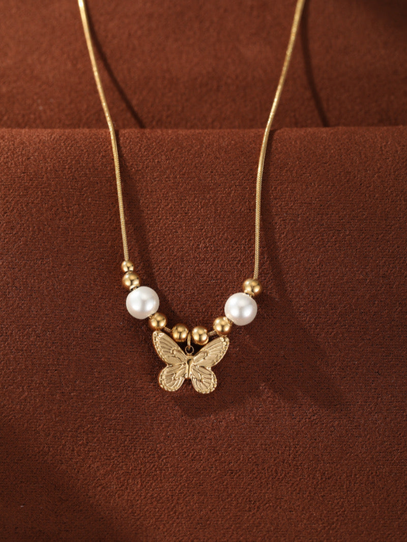Collier ton or présentant un pendentif papillon texturé suspendu entre deux perles blanches d'eau douce et de petites perles dorées sur une délicate chaîne serpent.