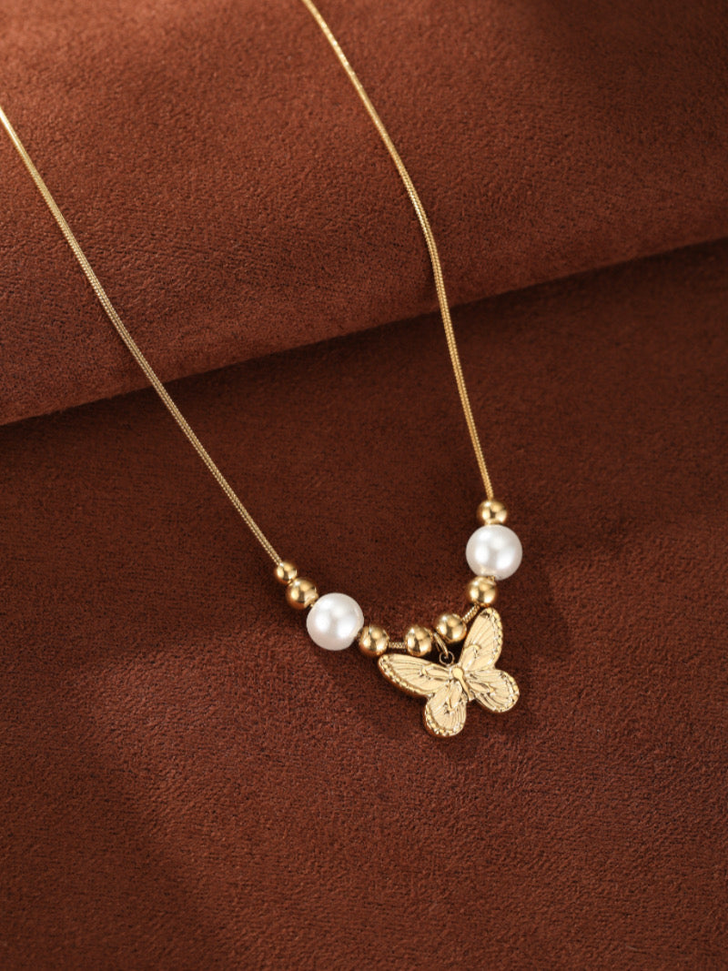Un délicat collier doré présentant un pendentif papillon détaillé, accentué de deux lustrées perles blanches d'eau douce et de petites perles dorées, présenté sur fond texturé marron chaleureux.